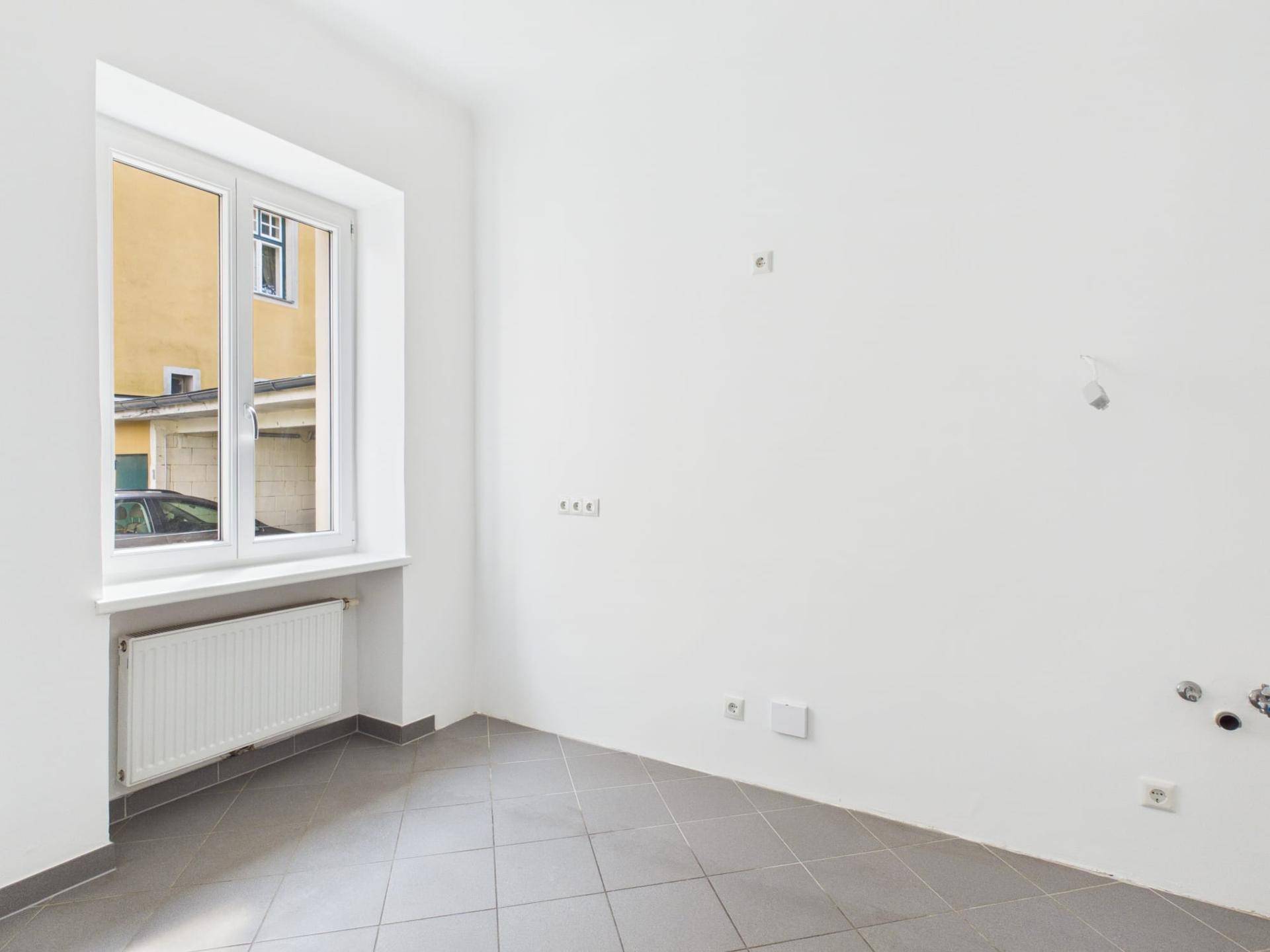 Heller Raum mit grauen Fliesen, Fenster und Anschlüssen für eine Küche.