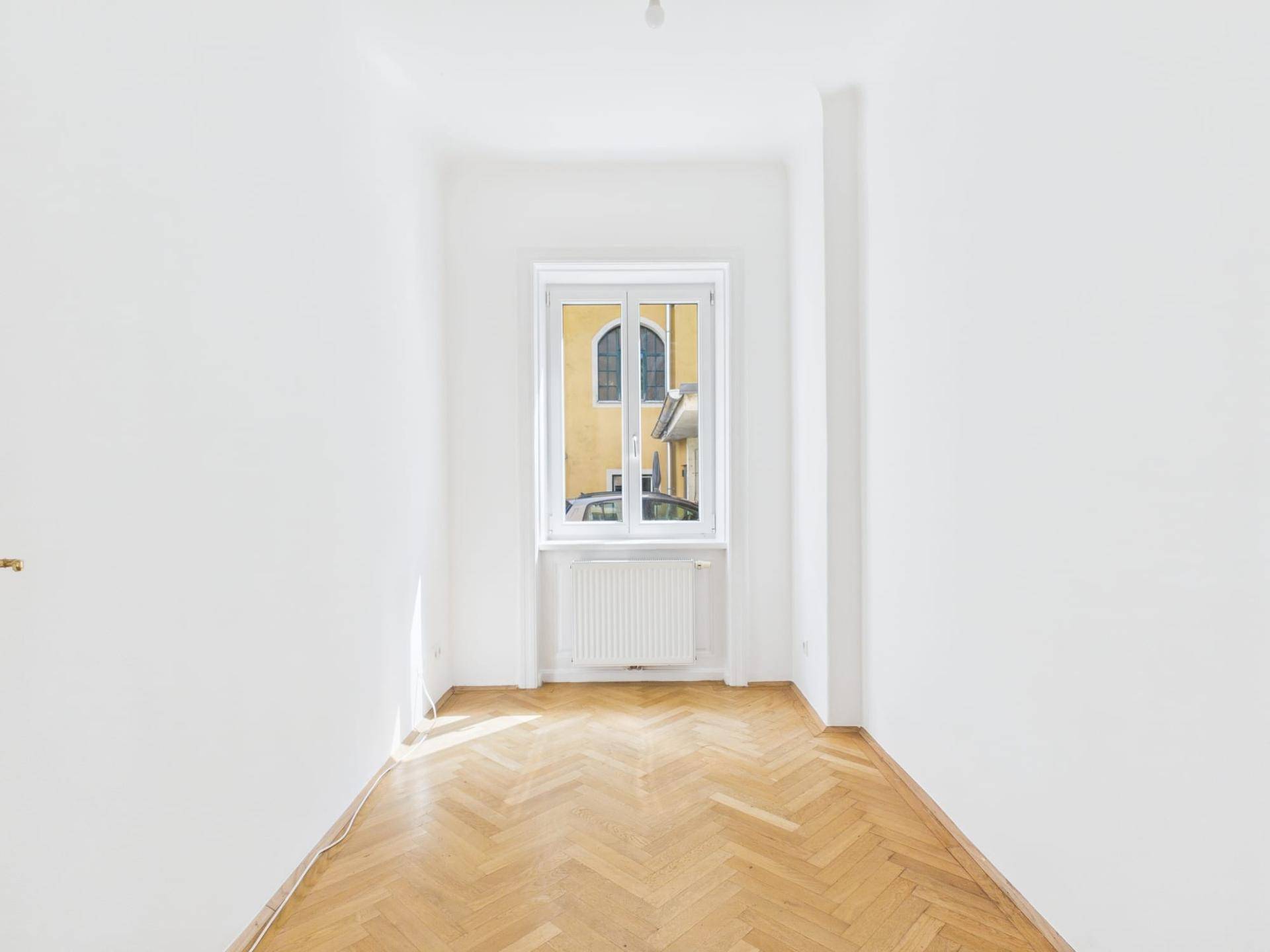 Helles Zimmer mit Fischgrätparkettboden, weißen Wänden und Blick in den Innenhof.