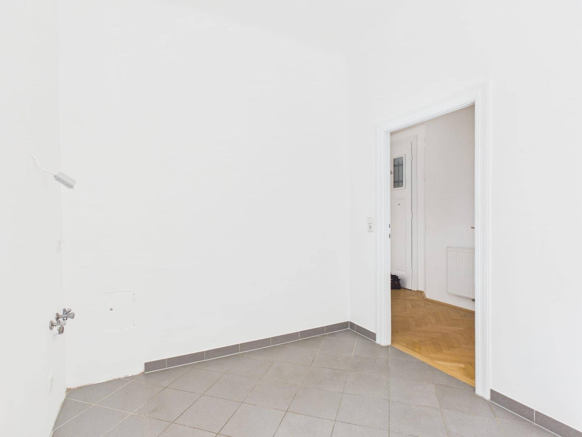 Leerer Raum mit grauen Fliesen und vorbereiteten Anschlüssen, angrenzend an einen Parkettraum.