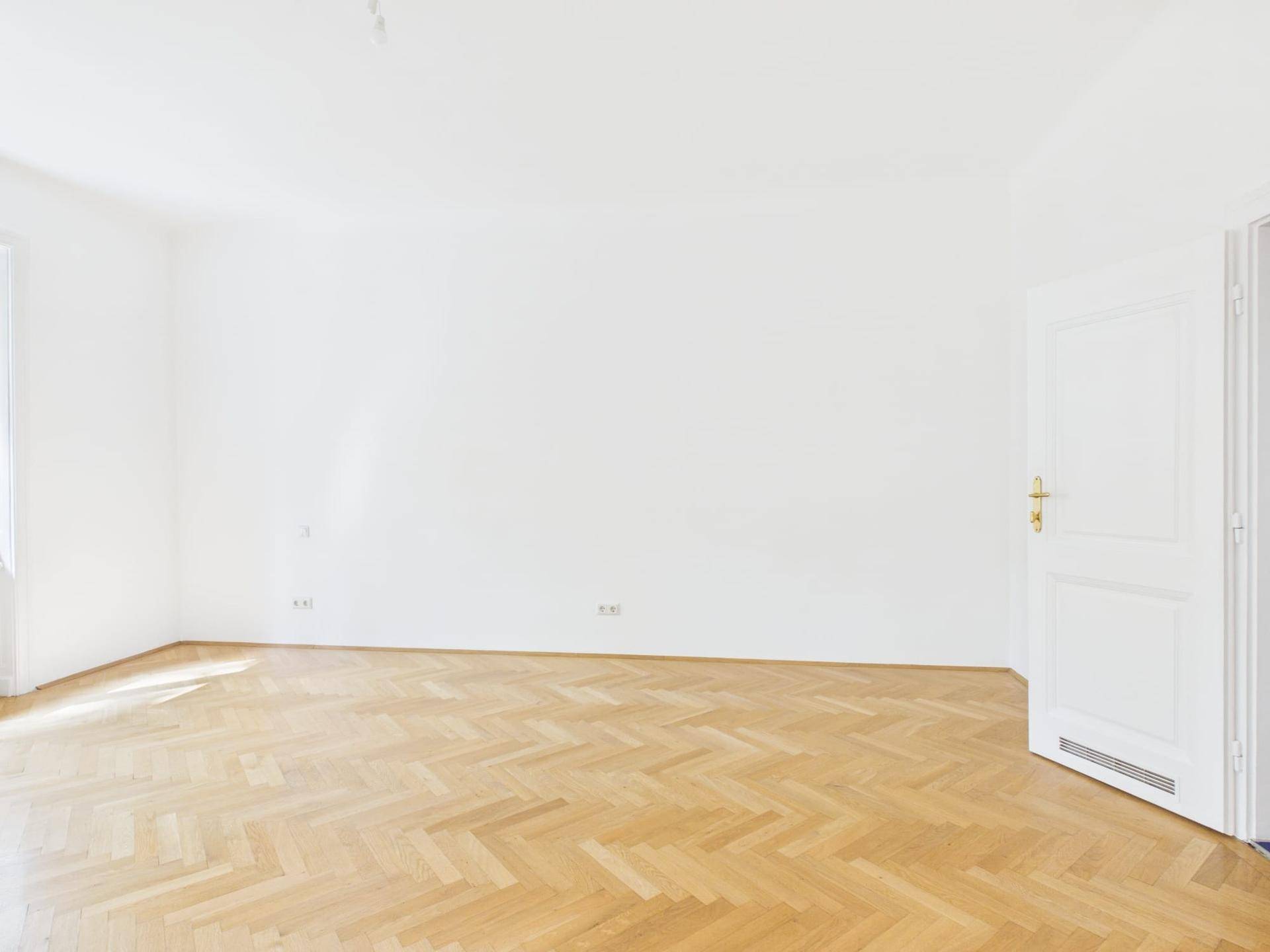 Geräumiges Zimmer mit hellem Fischgrätparkettboden und weißen Wänden.