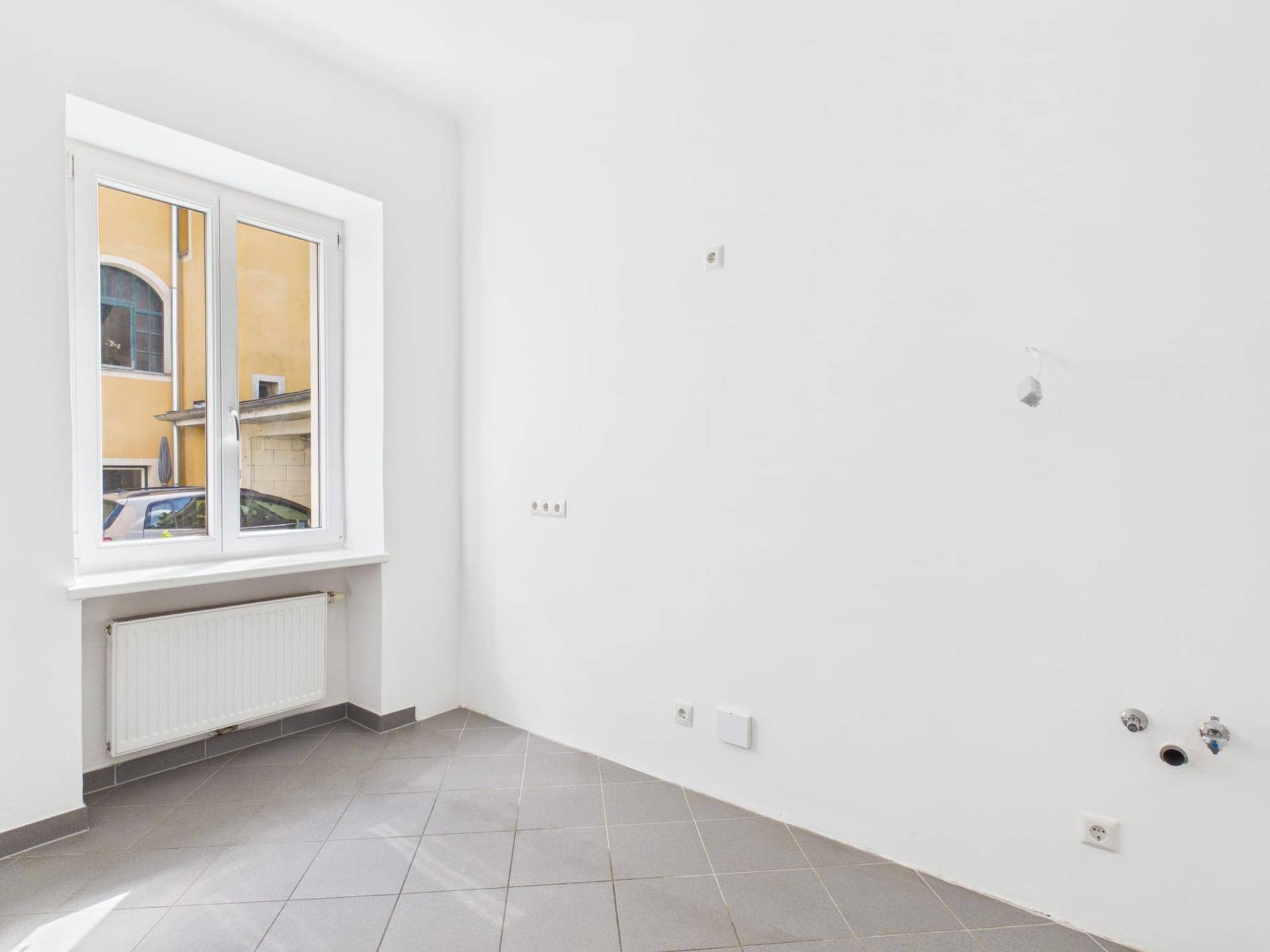 Heller Raum mit grauen Fliesen, Fenster und Anschlüssen für eine Küche.