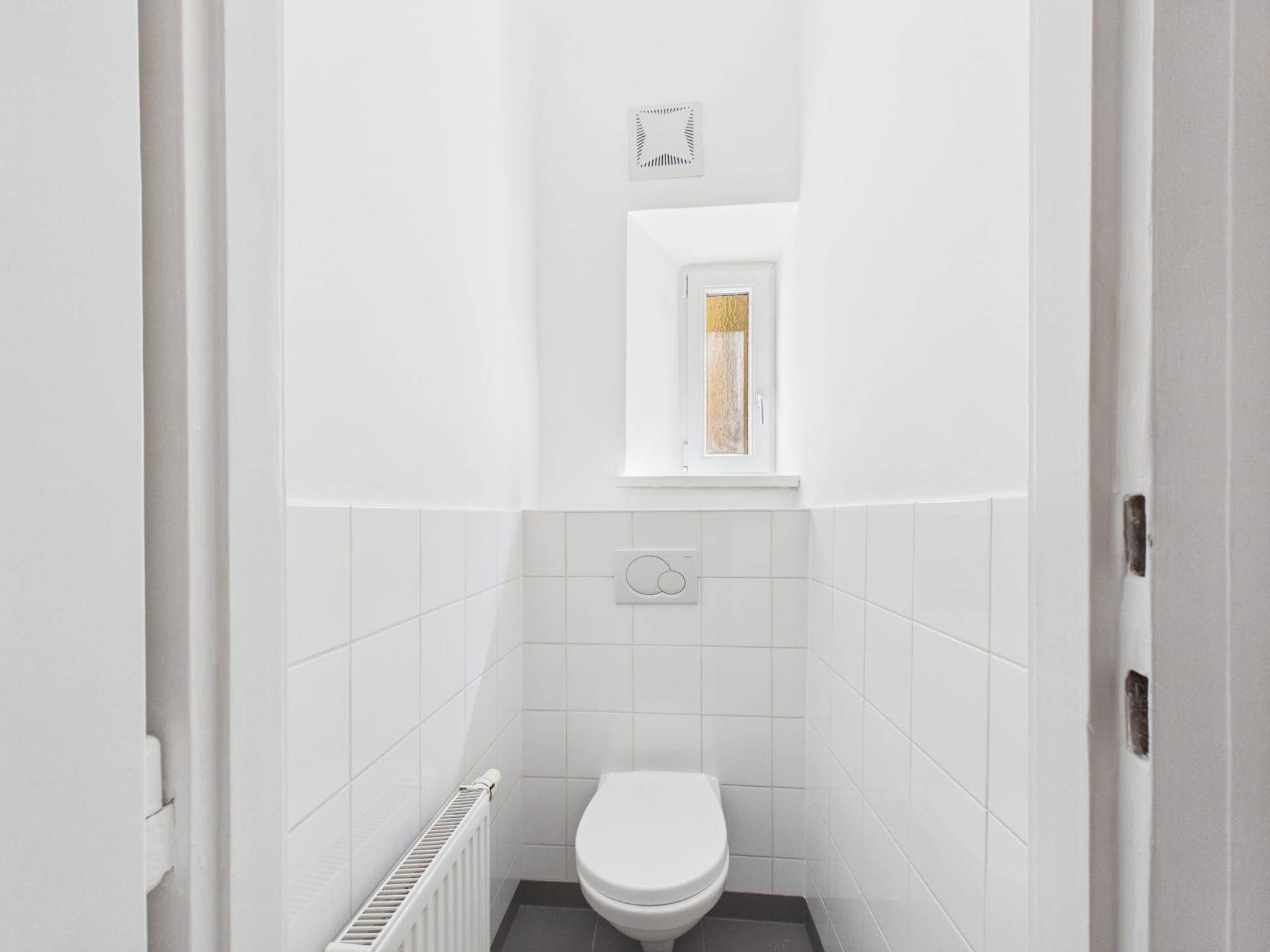 Kleines WC mit weißen Fliesen, wandhängender Toilette und Fenster für Tageslicht.