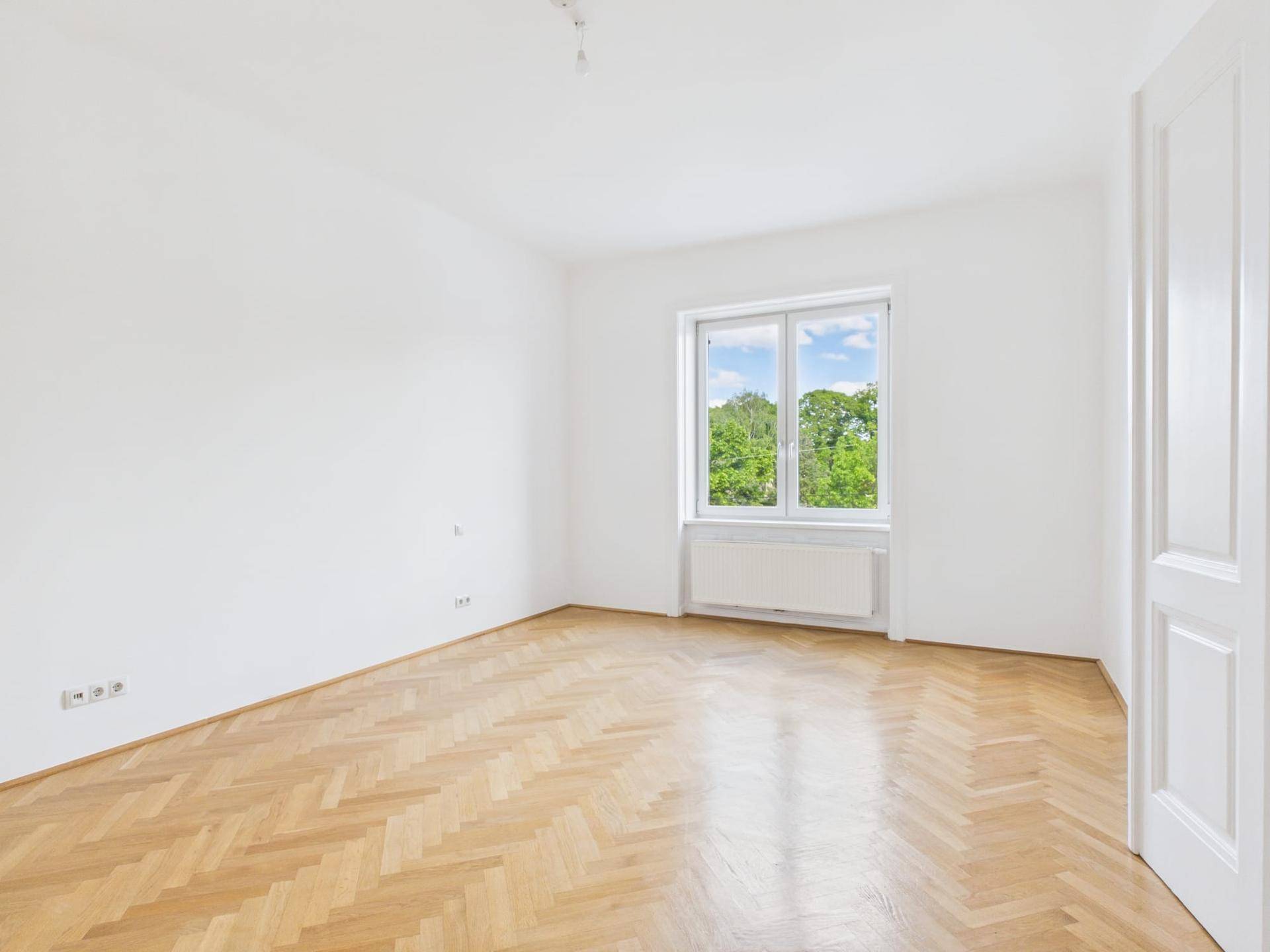 Leeres Zimmer mit hellem Fischgrätparkett, weißen Wänden und einem Fenster mit Blick ins Grüne.