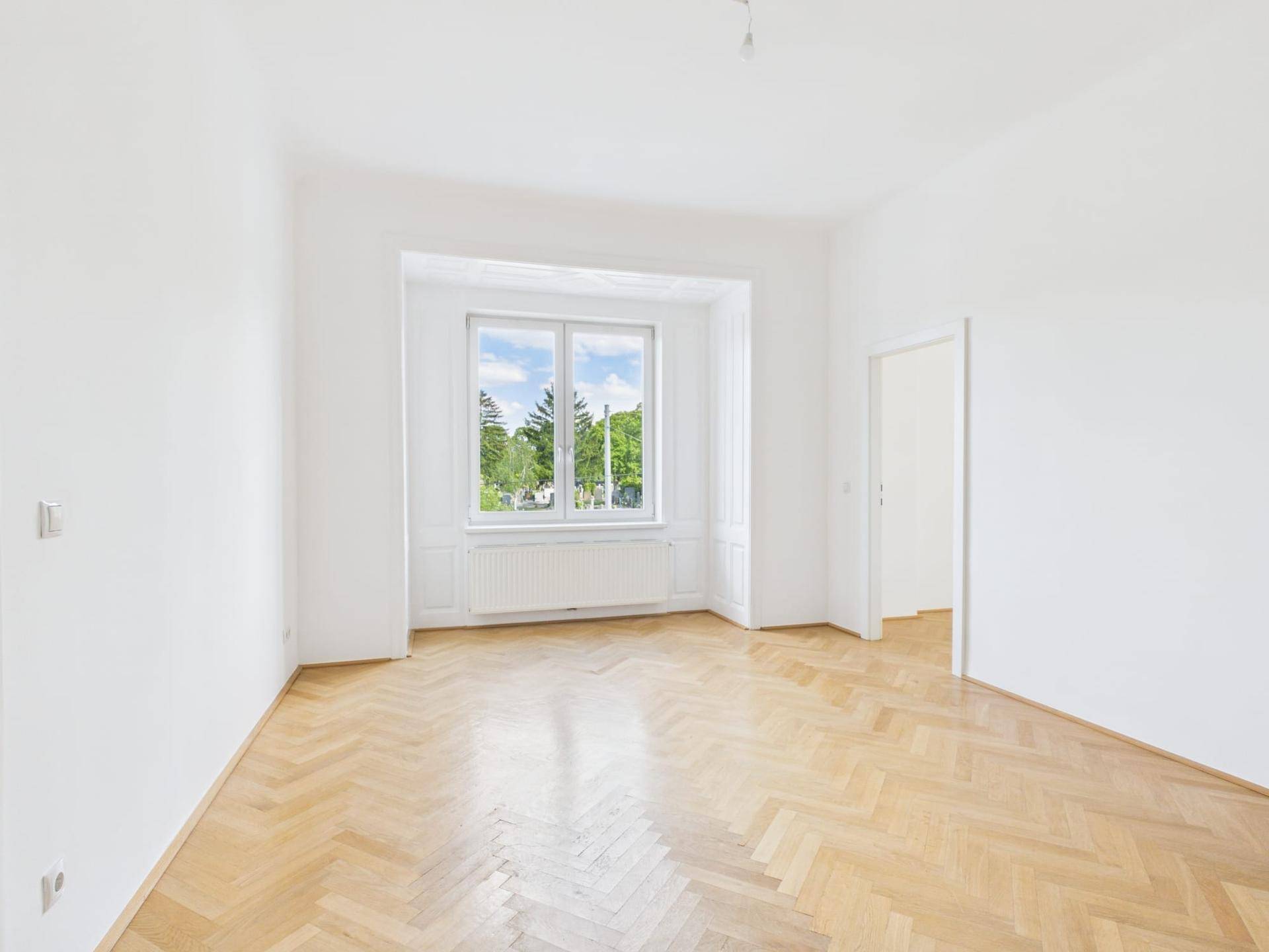 Geräumiges Zimmer mit Fischgrätparkett, hohen Decken und einem großen Fenster mit Blick ins Grüne.