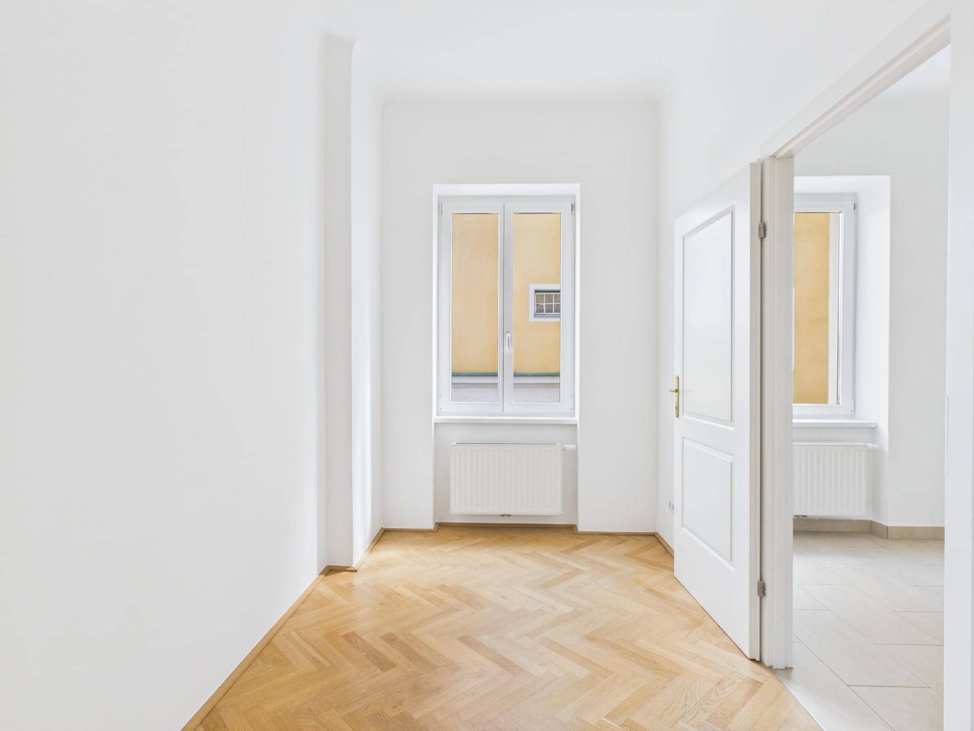 Helles Zimmer mit Fischgrätparkett und einem Fenster, das den Blick auf eine gegenüberliegende Fassade freigibt.