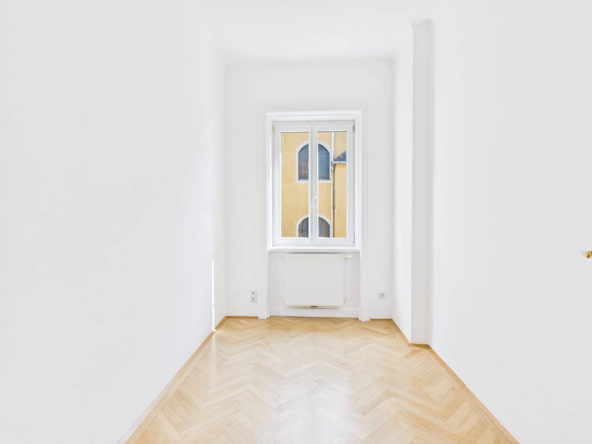 Schmaler Raum mit Fischgrätparkett und einem Fenster, das auf ein gegenüberliegendes Gebäude blickt.