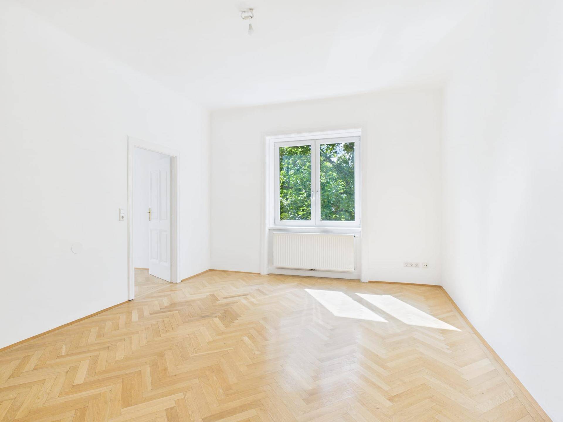 Geräumiges, helles Zimmer mit Fischgrätparkett und Fenster mit Blick auf Bäume.