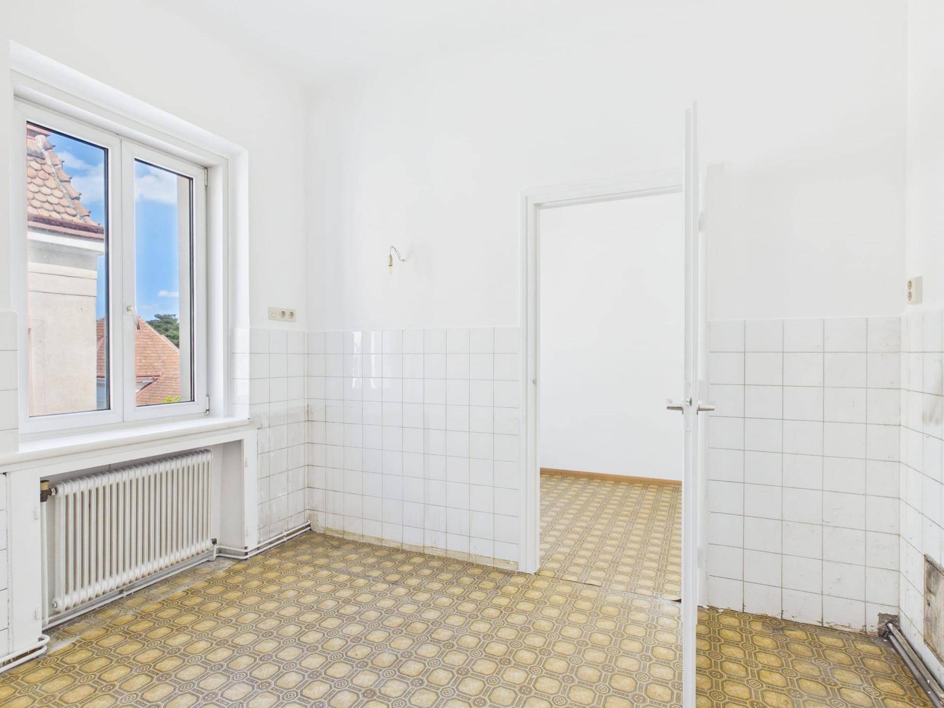 Küche mit weißen Wandfliesen, gemustertem Boden und einem Fenster mit Blick auf die Nachbarschaft.