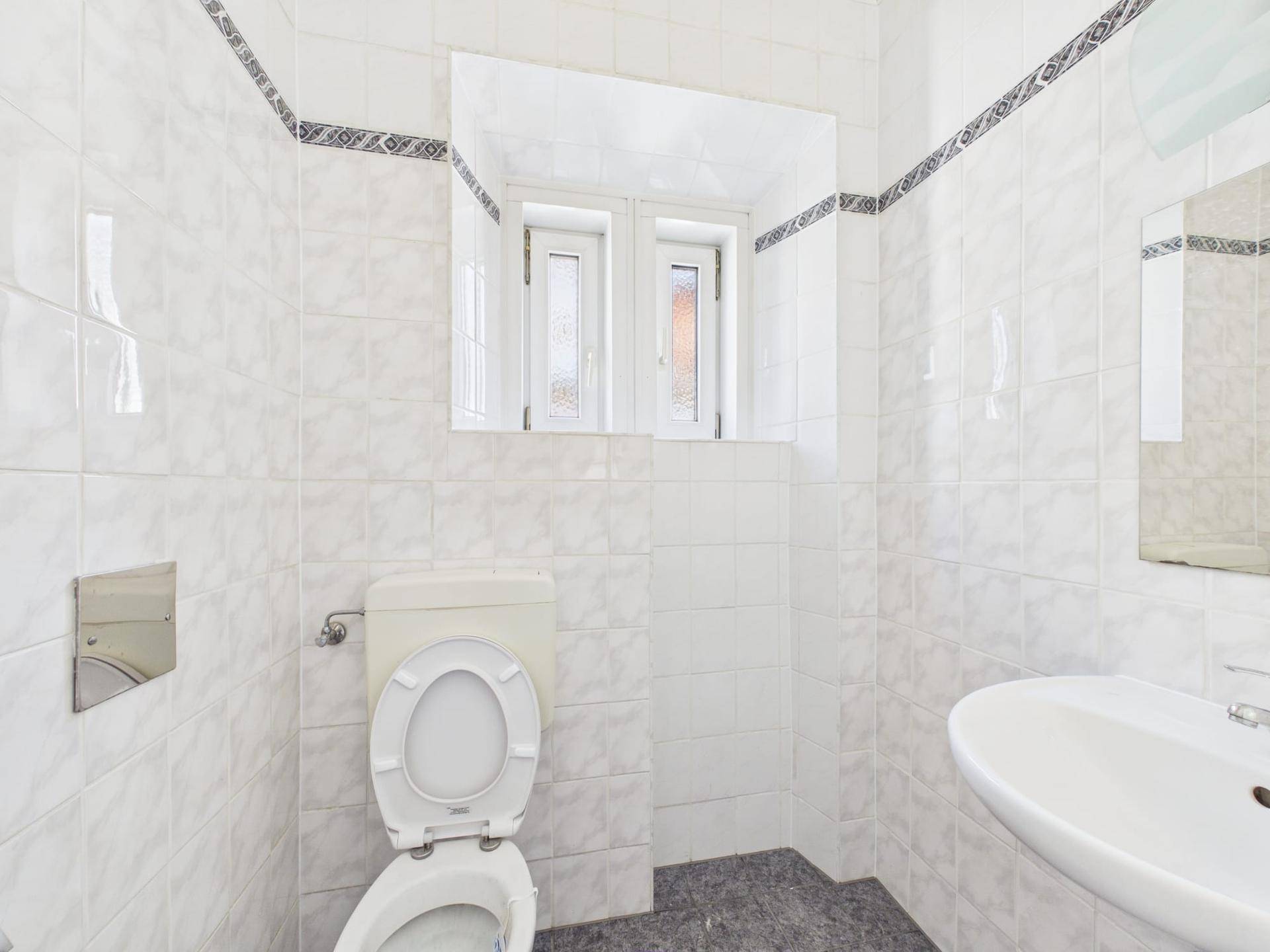 WC-Bereich mit weißen Fliesen, Toilette, Waschbecken und Fenster für natürliche Belüftung.