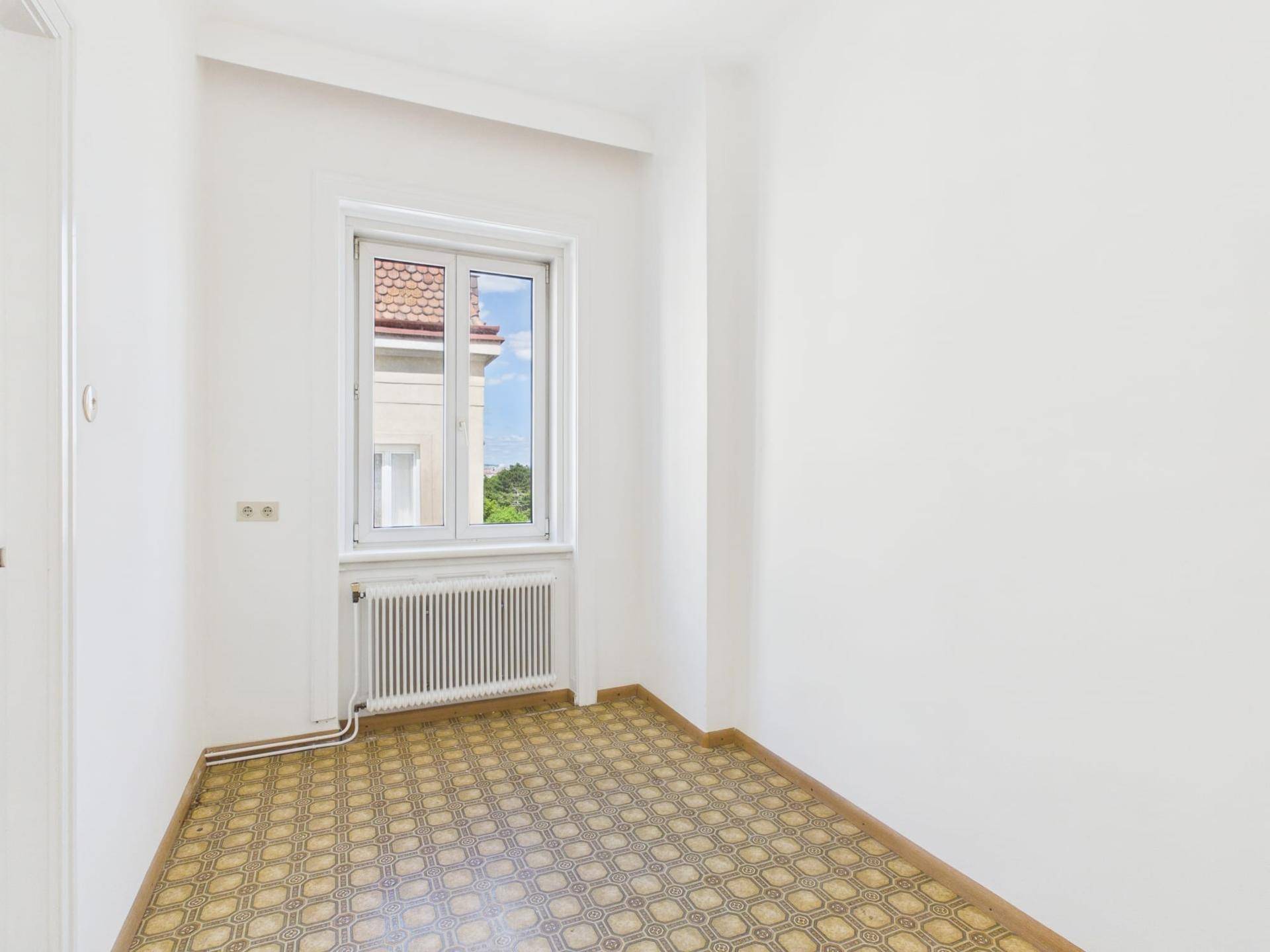 Kleiner Raum mit gemustertem Boden, weiß gestrichenen Wänden und Fensterblick auf ein Nachbargebäude.