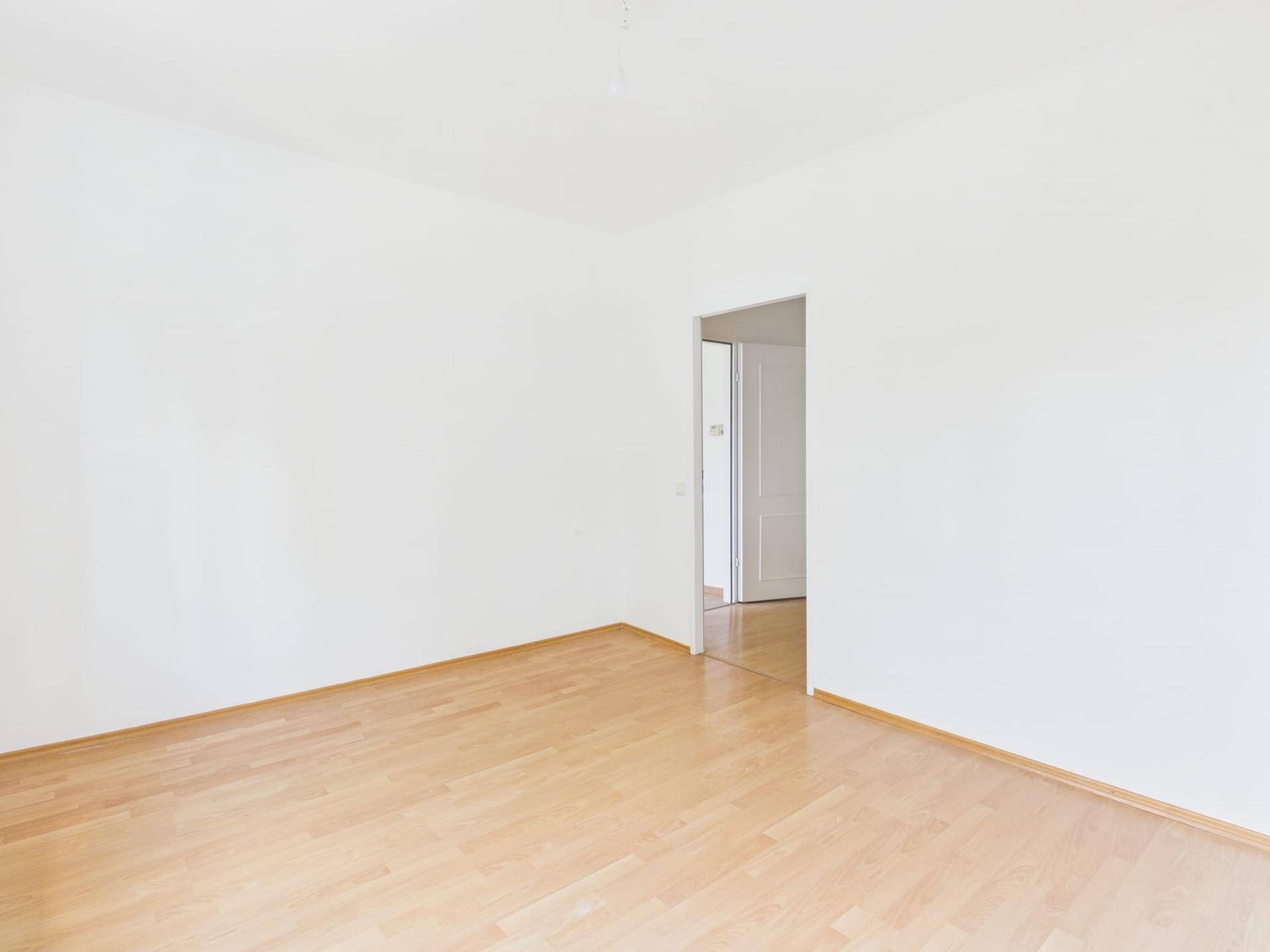 Leerer Raum mit Laminatboden und weiß gestrichenen Wänden, mit Zugang zu einem weiteren Zimmer.