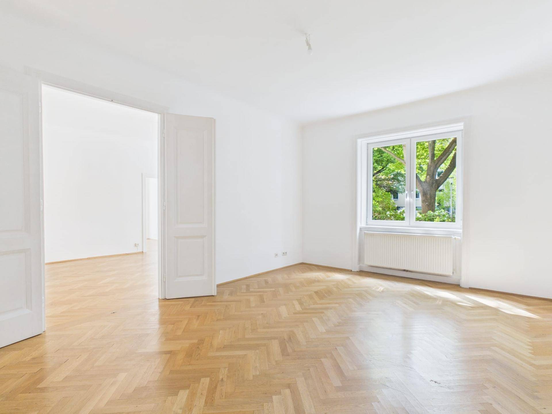 Geräumiges, helles Zimmer mit Fischgrätparkett und Blick ins Grüne durch das Fenster.