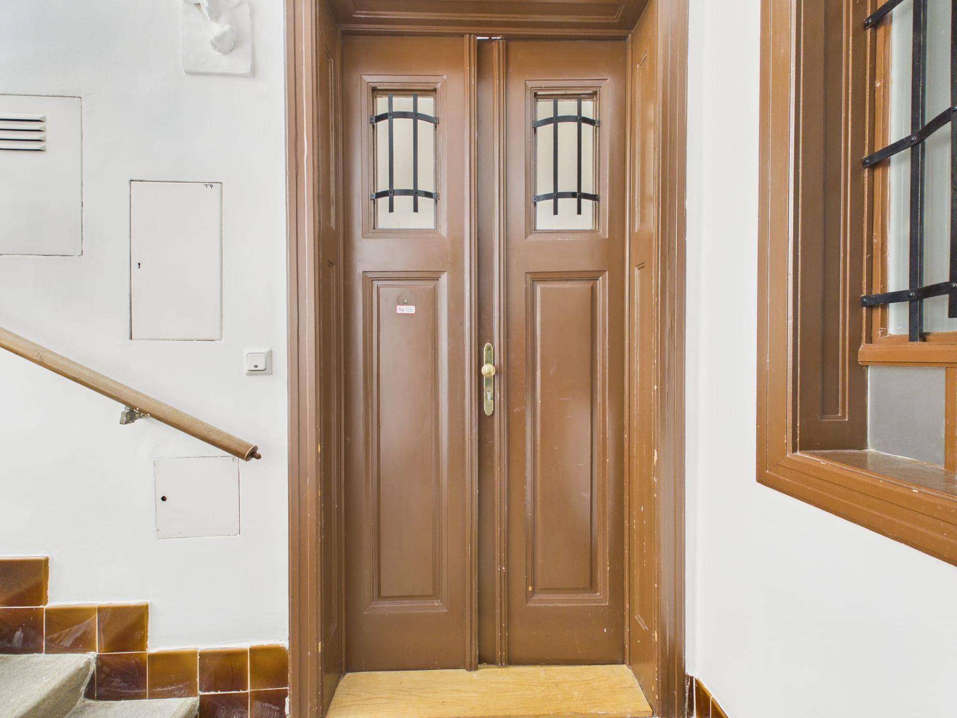 Braune Eingangstür mit dekorativen Gittern in einem hellen Treppenhaus mit Fliesenboden.