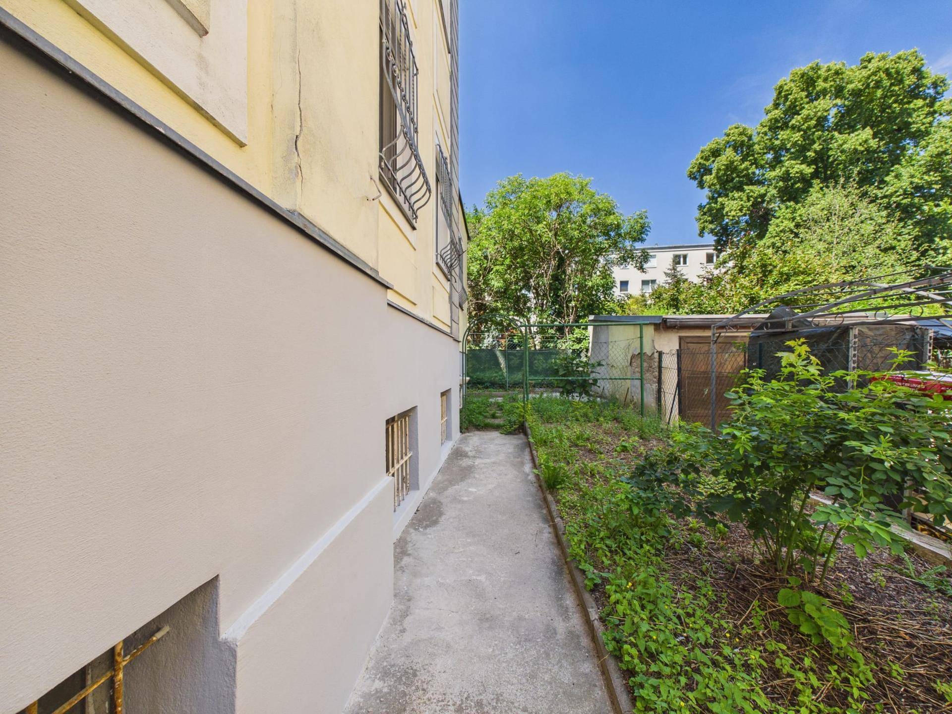 Schmaler Weg entlang der Gebäudeseite mit Bepflanzung und Blick auf den Garten.