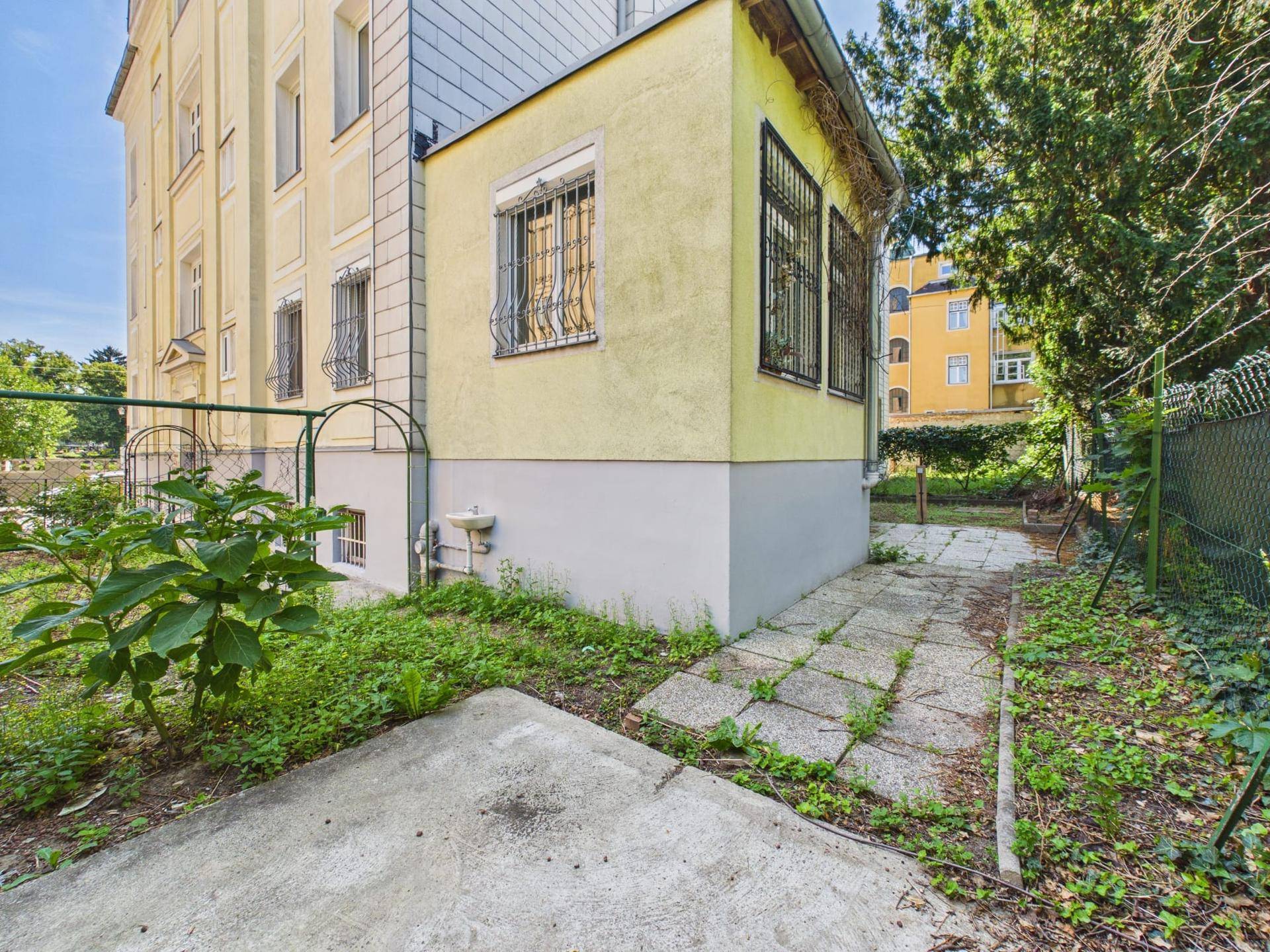 Seitenansicht des Gebäudes mit einem kleinen, ungepflegten Gartenbereich und gepflastertem Weg.