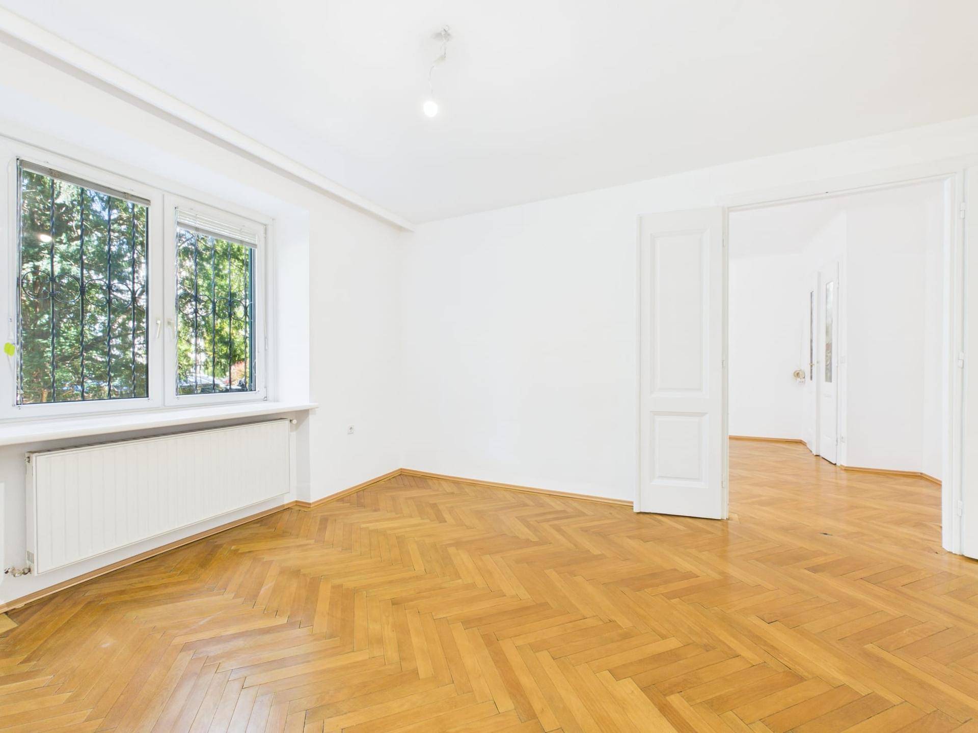 Geräumiges Wohnzimmer mit Fischgrätparkett, weißen Wänden und viel Tageslicht.