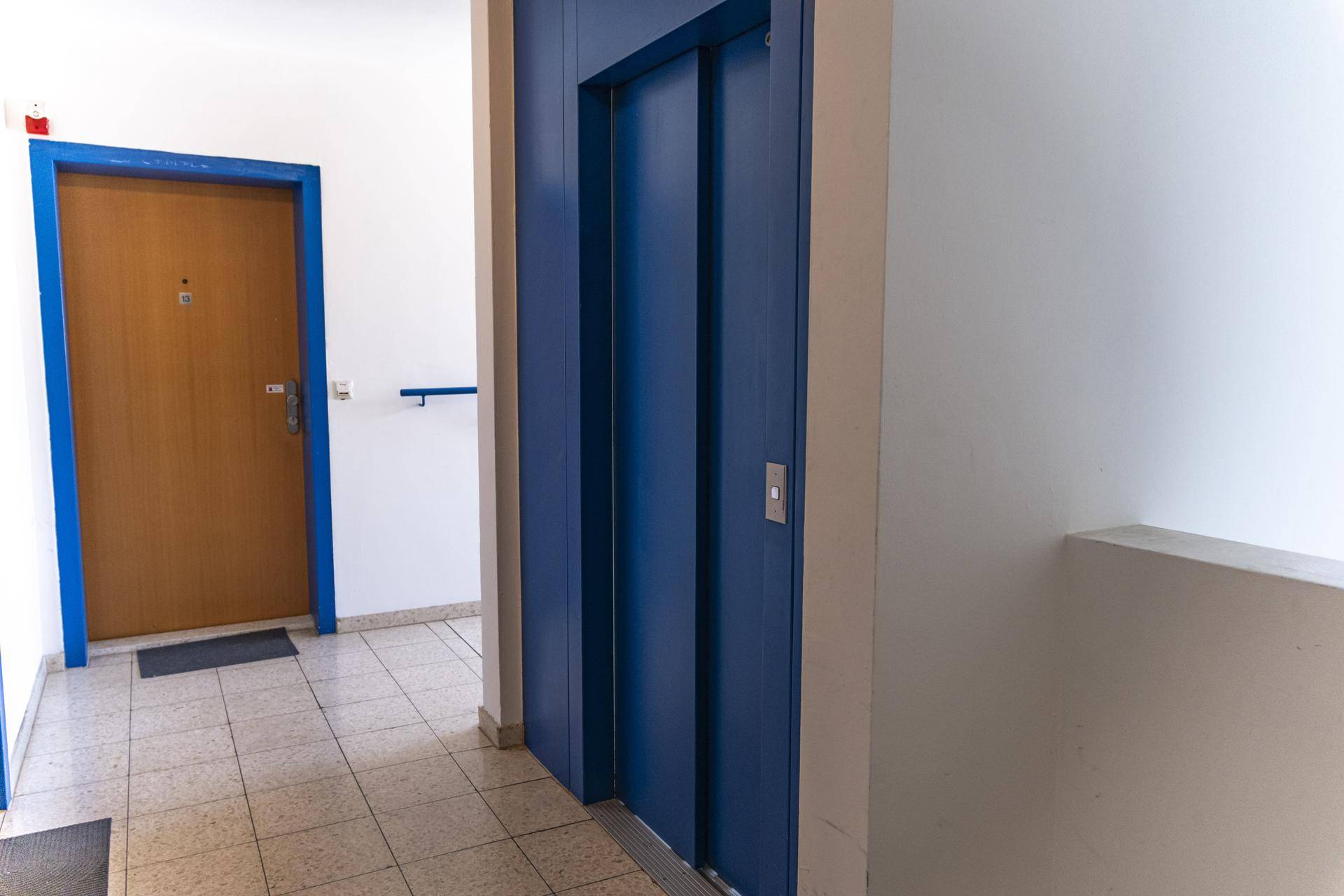 Heller Flur mit blauem Aufzug und einer Holztür zu einer Wohnung.