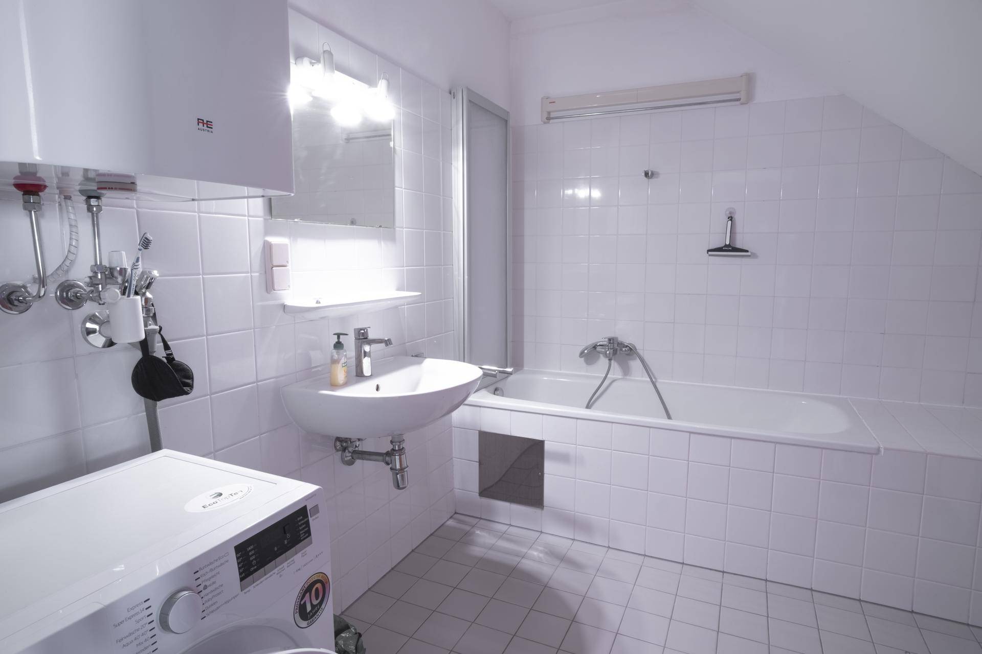 Helles Badezimmer mit Badewanne, Waschbecken und Waschmaschine, komplett gefliest in Weiß.
