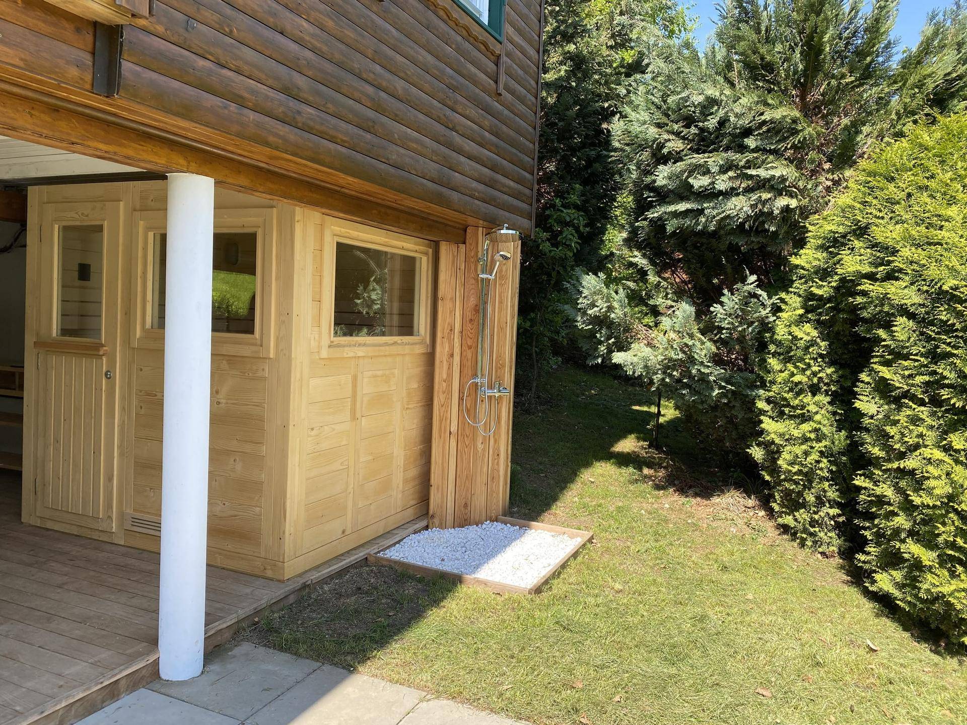 Sauna-Hütte und Außendusche im Garten, umgeben von üppigem Grün und Bäumen.