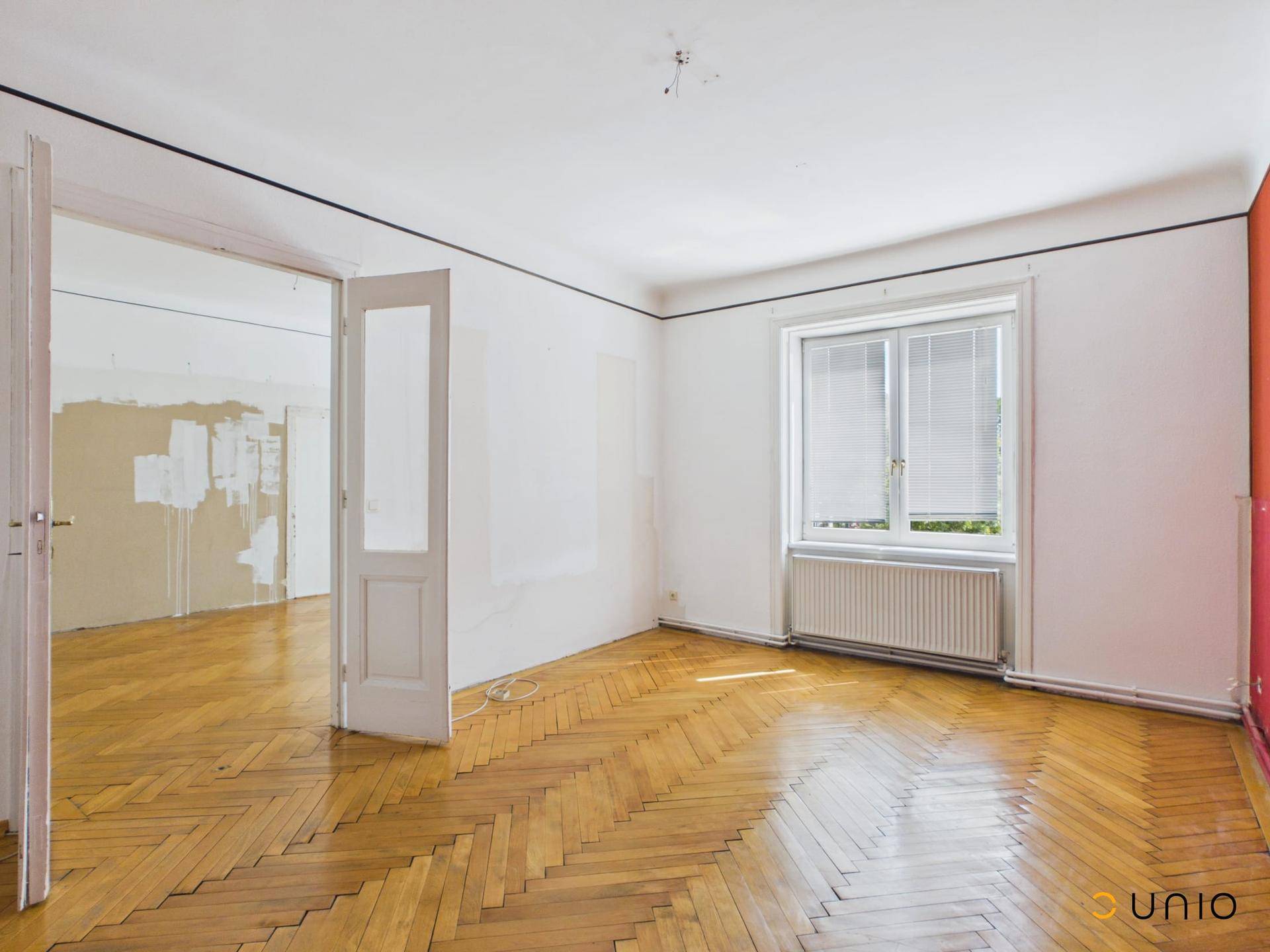 Großes Zimmer mit Fischgrätparkett und Fenster, das auf eine Renovierung wartet.