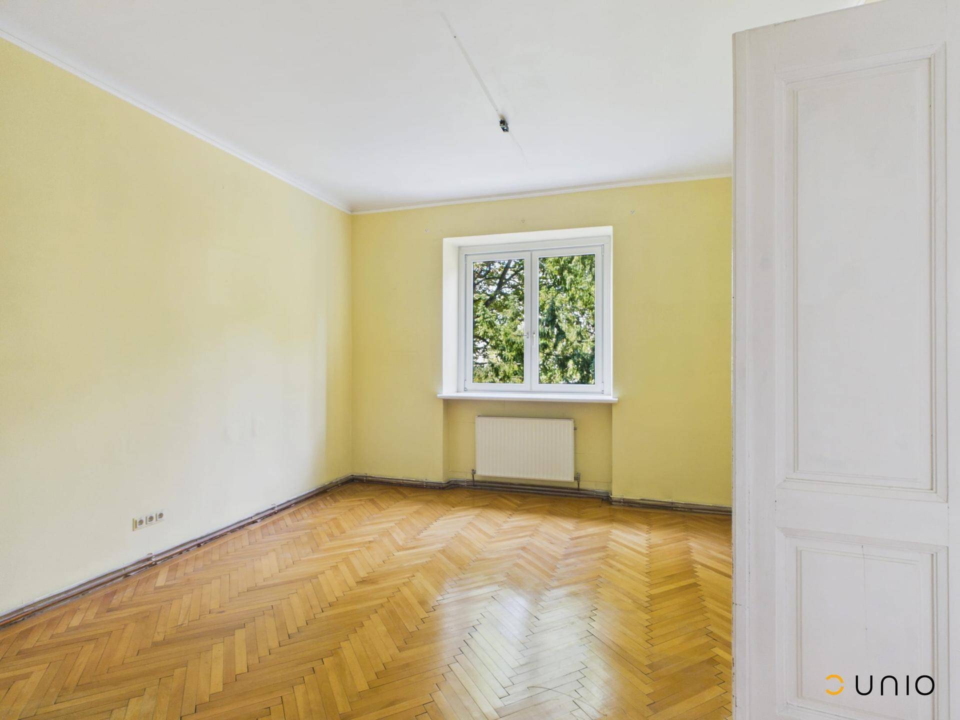 Geräumiges Zimmer mit gelben Wänden, Fischgrätparkett und einem Fenster mit Blick ins Grüne.