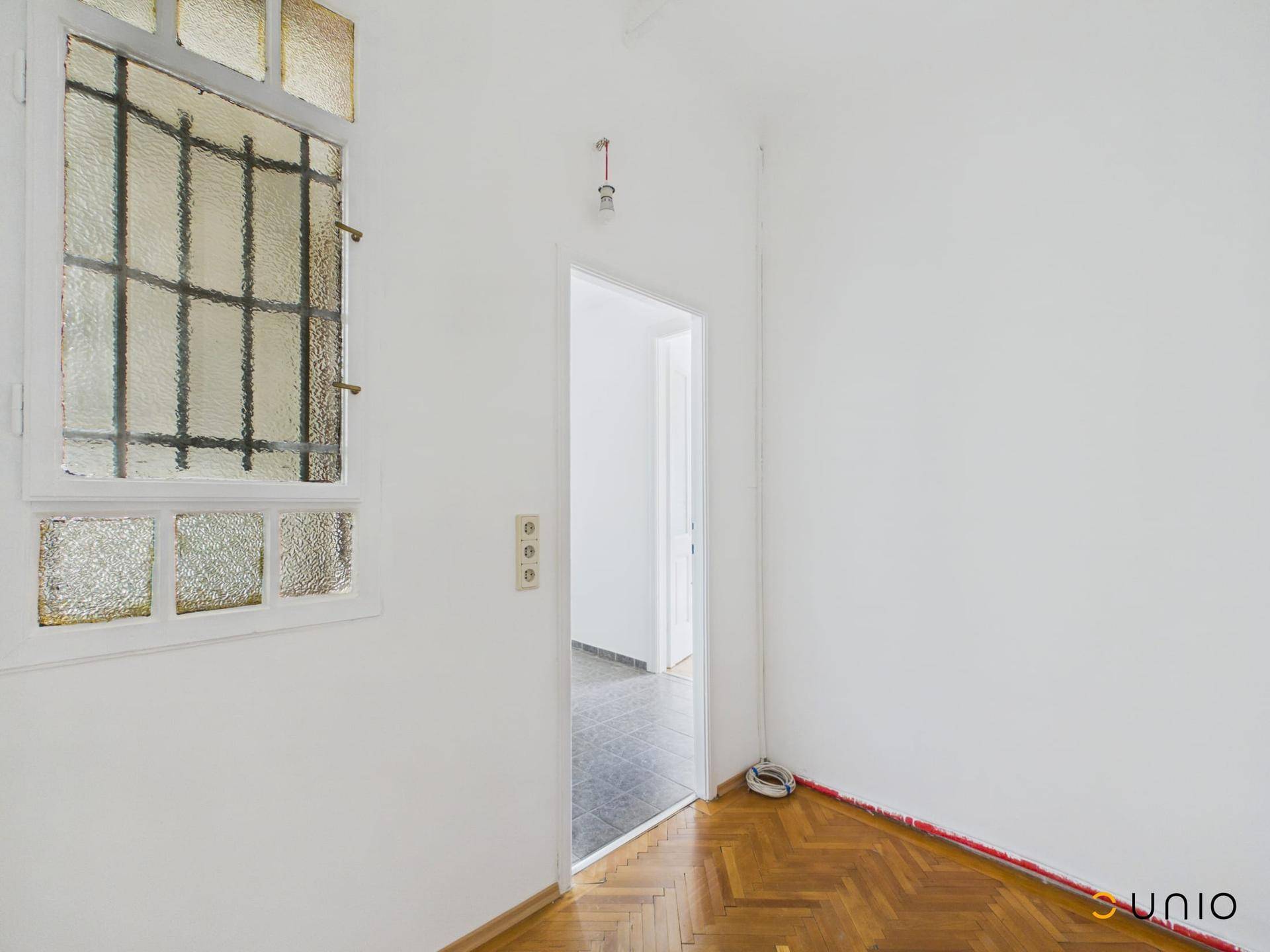 Ecke eines Zimmers mit Fischgrätparkett und einem Fenster mit Gitter.