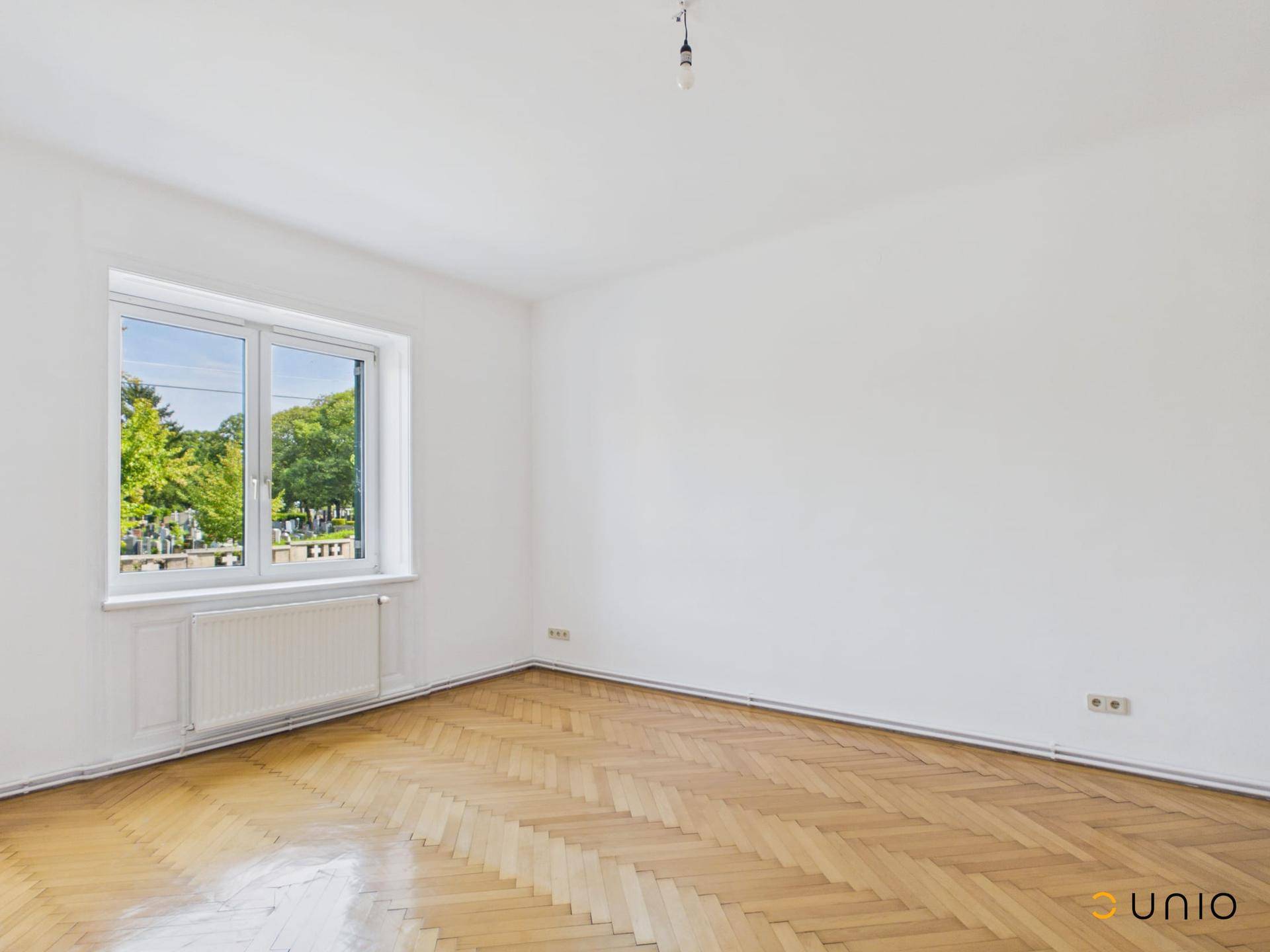 Helles Zimmer mit Fischgrätparkett und großem Fenster mit Blick ins Grüne.
