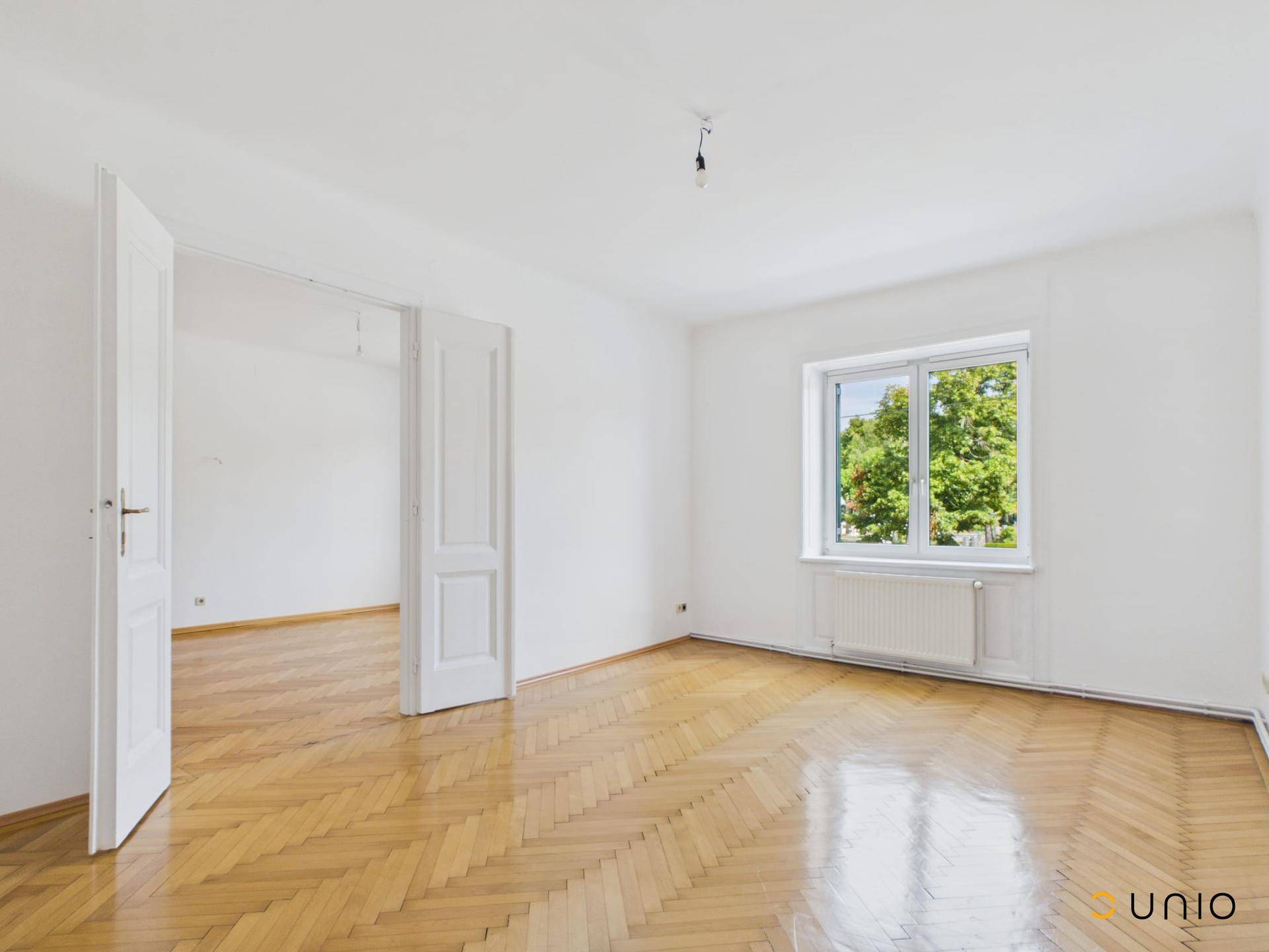 Großes Zimmer mit Fischgrätparkett und Zugang zu einem weiteren Raum, ideal als Wohnzimmer.