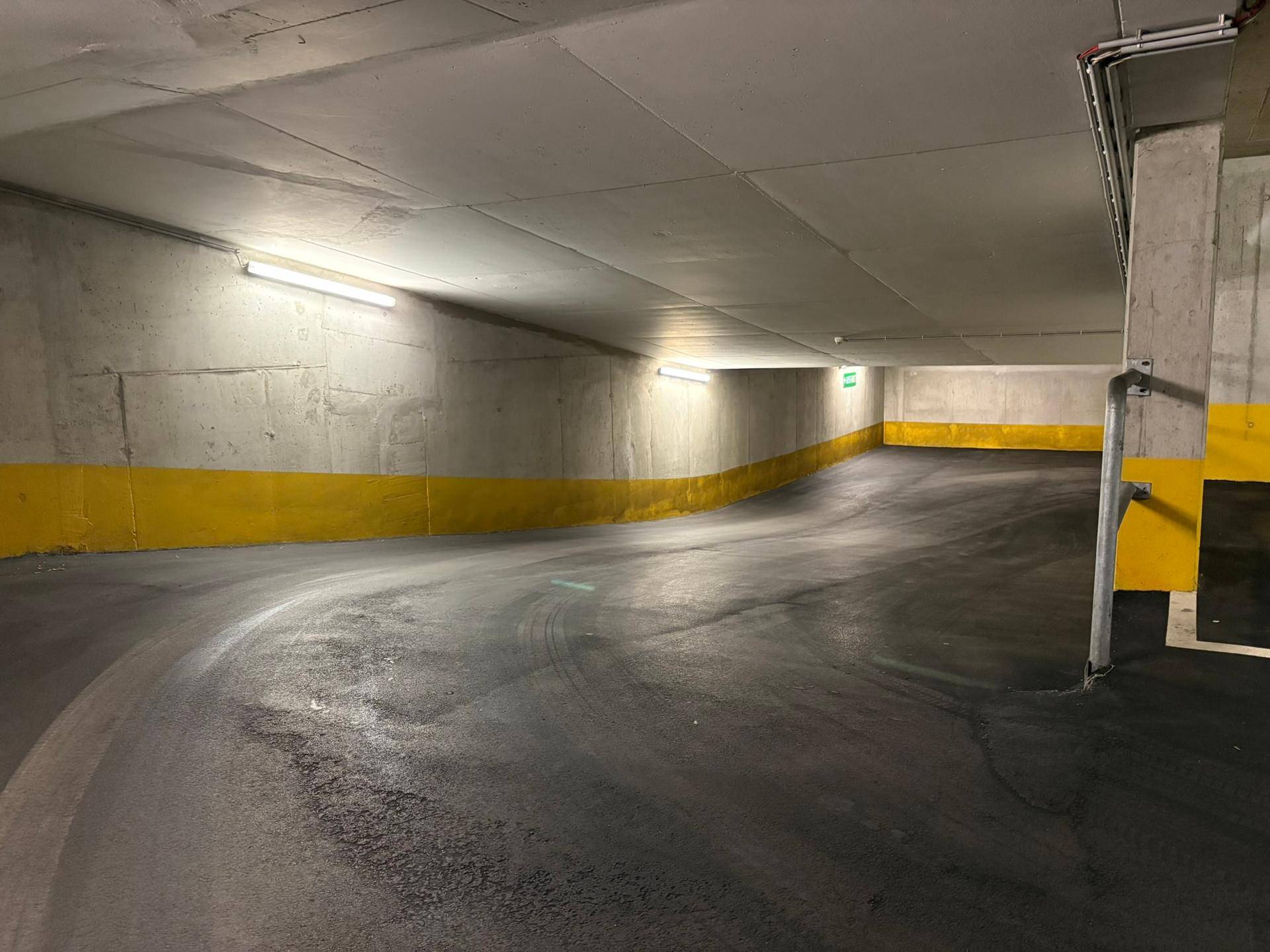 Zufahrtsrampe in einer Tiefgarage mit Betonwänden, gelber Markierung und heller Beleuchtung.