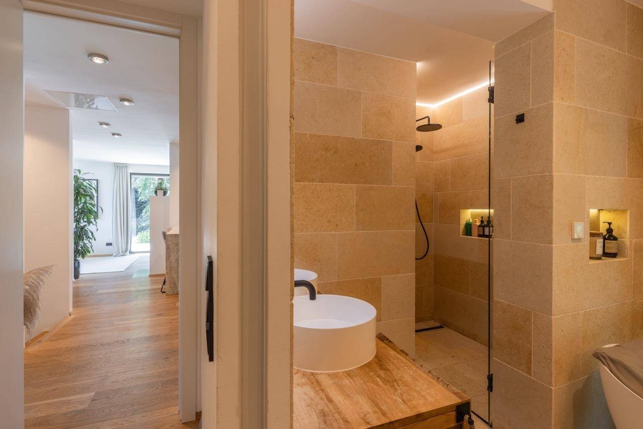 Elegantes Badezimmer mit begehbarer Dusche, Natursteinfliesen und Blick in den Flur.
