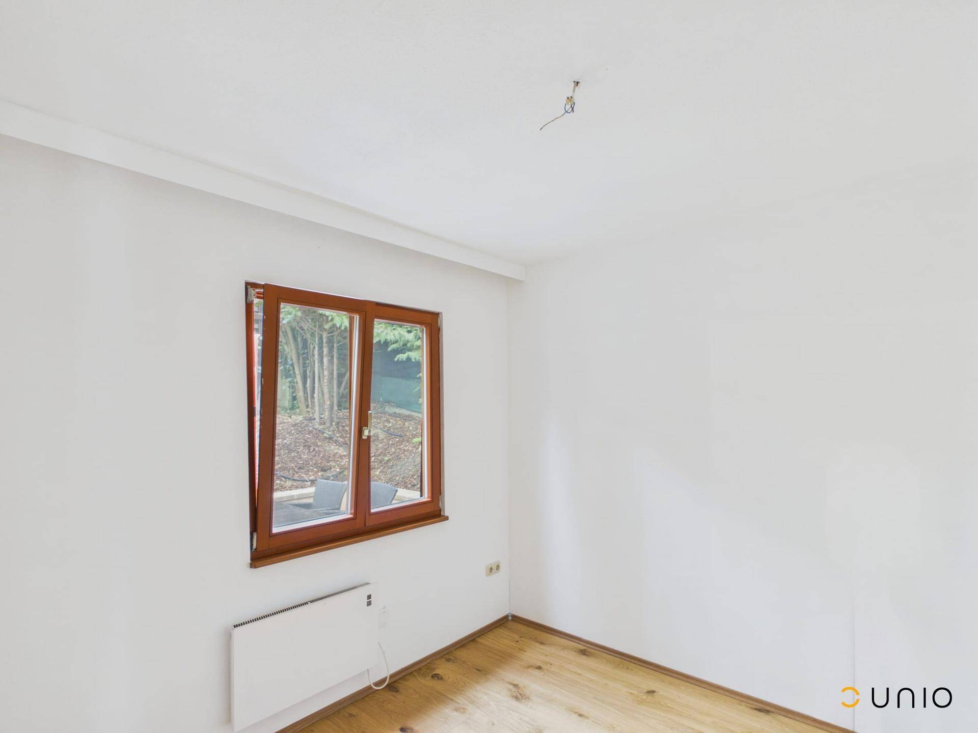 Heller Raum mit Holzboden, weißen Wänden und einem Fenster mit Blick auf die Bepflanzung.