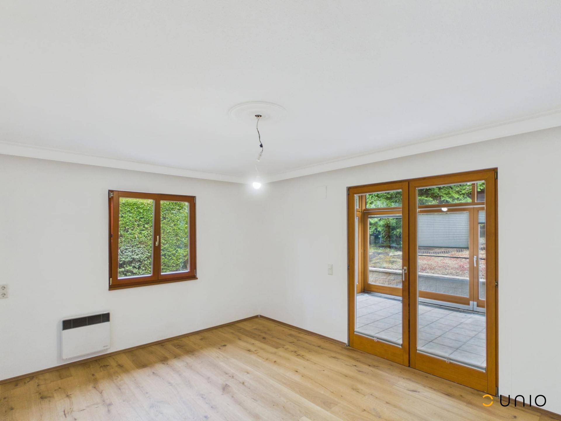 Leeres Wohnzimmer mit Holzboden, weißen Wänden und Zugang zu einem Wintergarten.