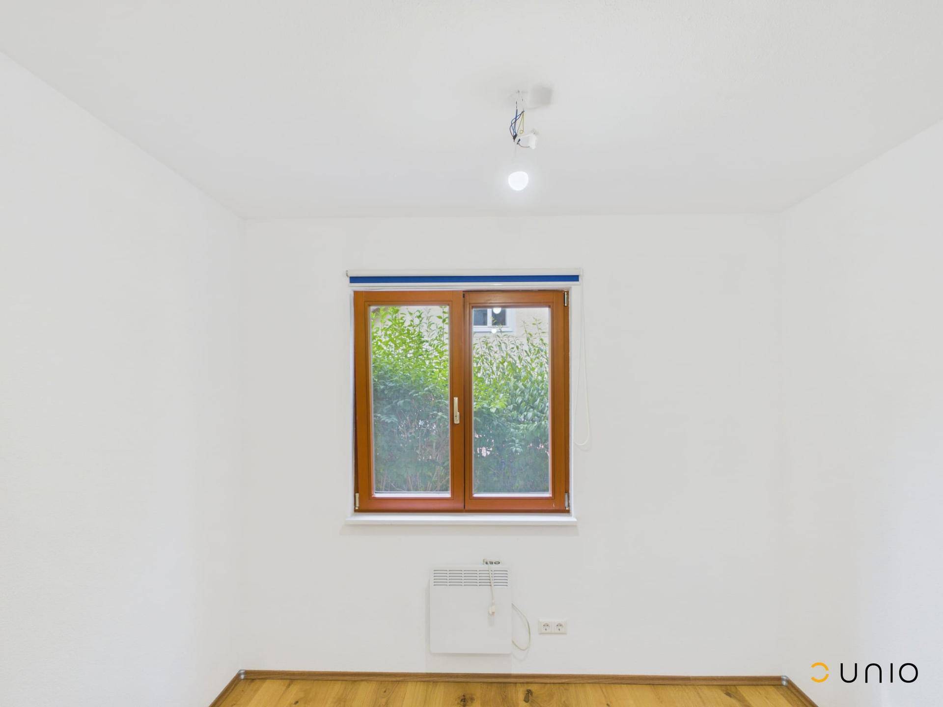 Heller Raum mit Holzboden, weiß gestrichenen Wänden und einem Fenster mit Blick ins Grüne.