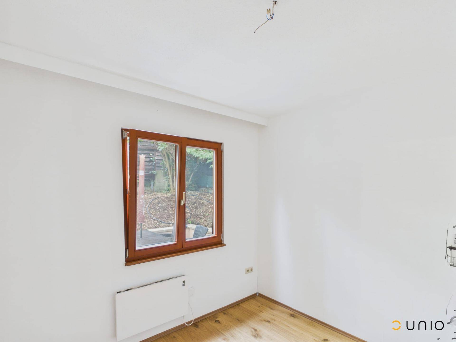 Leerer Raum mit Holzboden, weißen Wänden und einem Fenster mit Blick auf die Außenanlage.