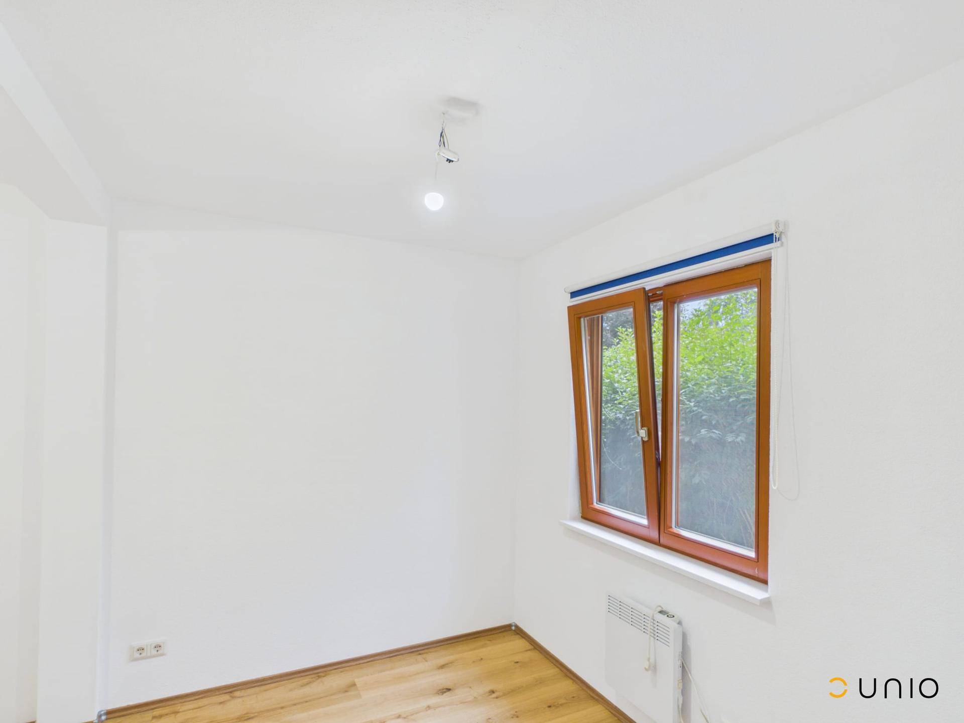 Leerer Raum mit Holzboden, weiß gestrichenen Wänden und einem Fenster mit Blick ins Grüne.