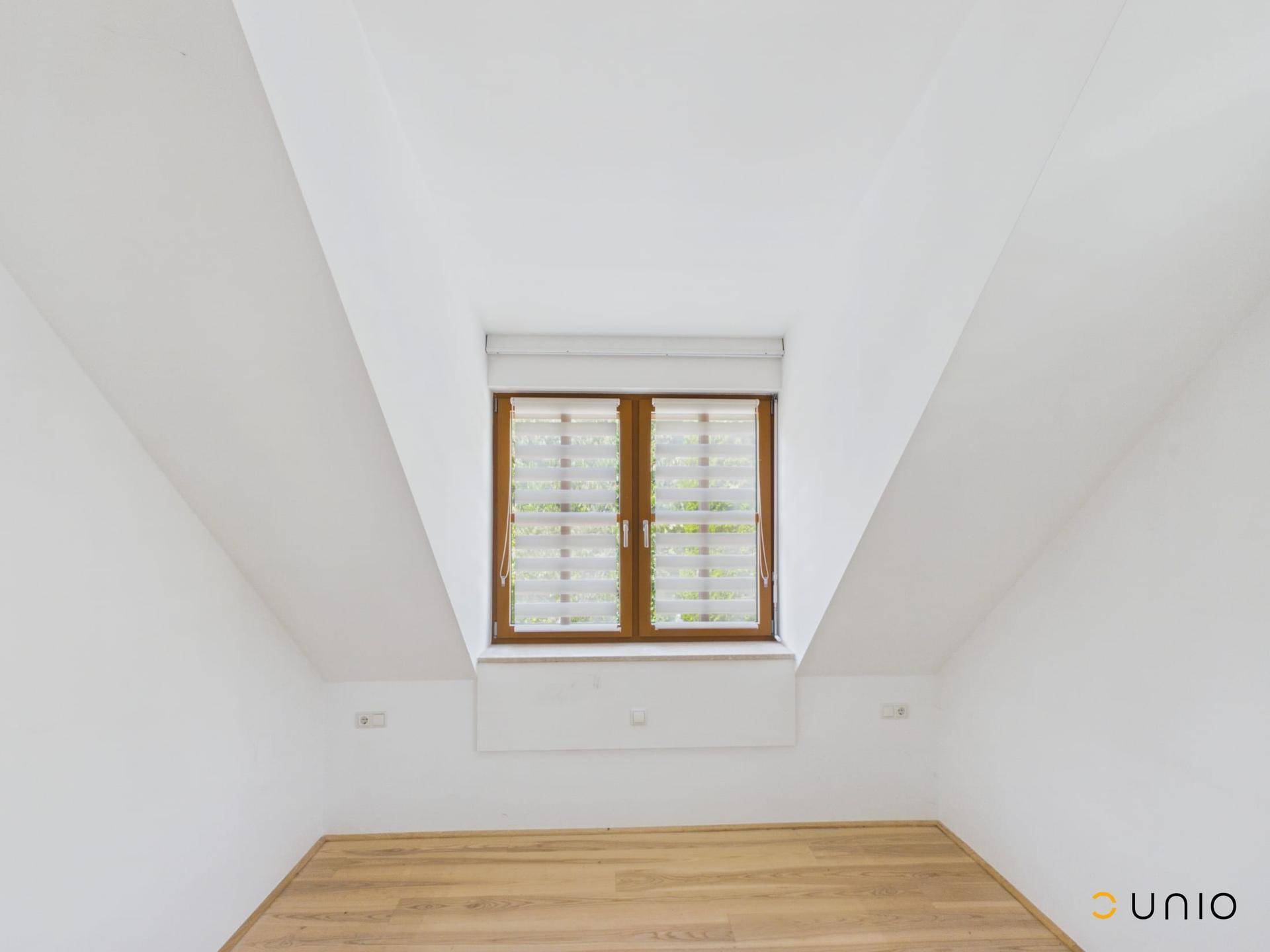 Helles Dachzimmer mit Holzfußboden und einem großen Fenster, das viel Tageslicht hereinlässt.