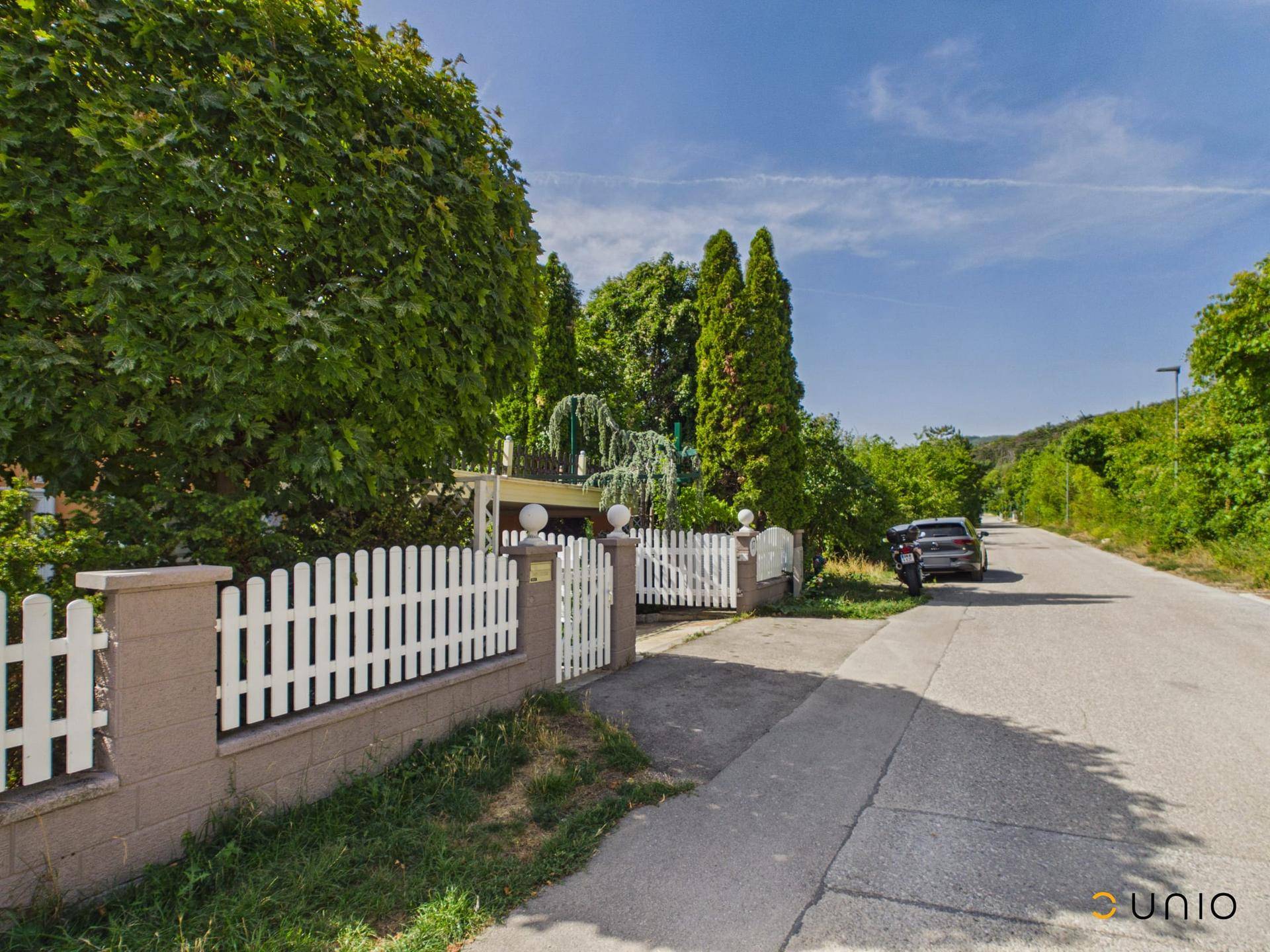 Ansicht des Hauses von der Straße mit weißem Zaun, Bäumen und einem gepflasterten Zugang.
