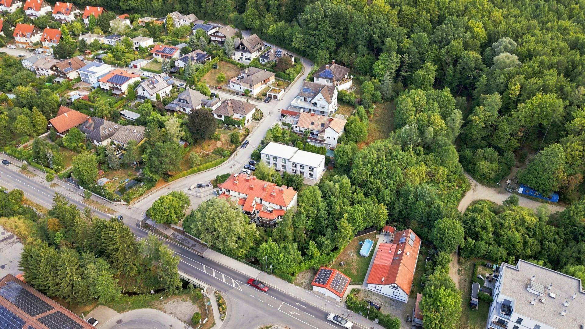 Luftaufnahme einer Wohnsiedlung mit viel Grün, Wald und einer Straße mit parkenden Autos.