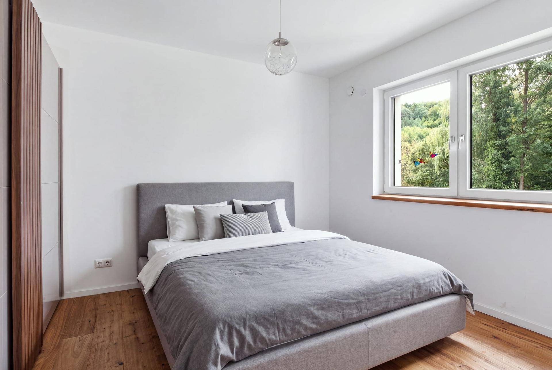 Helles Schlafzimmer mit Holzboden, großem Fenster und Blick in die Natur.