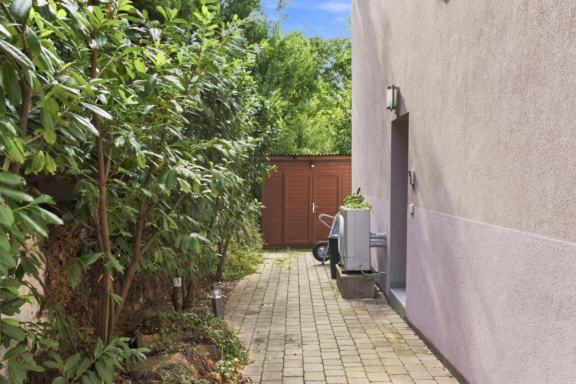 Gepflasterter Weg entlang des Hauses mit üppiger Bepflanzung und einem Gartenschuppen im Hintergrund.