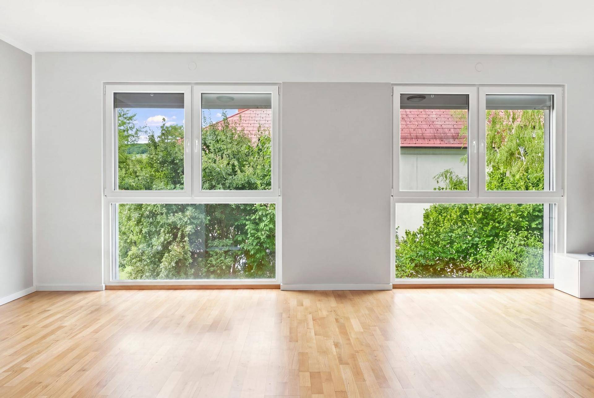 Leeres, helles Zimmer mit Holzboden und großen Fenstern, die viel Tageslicht hereinlassen.