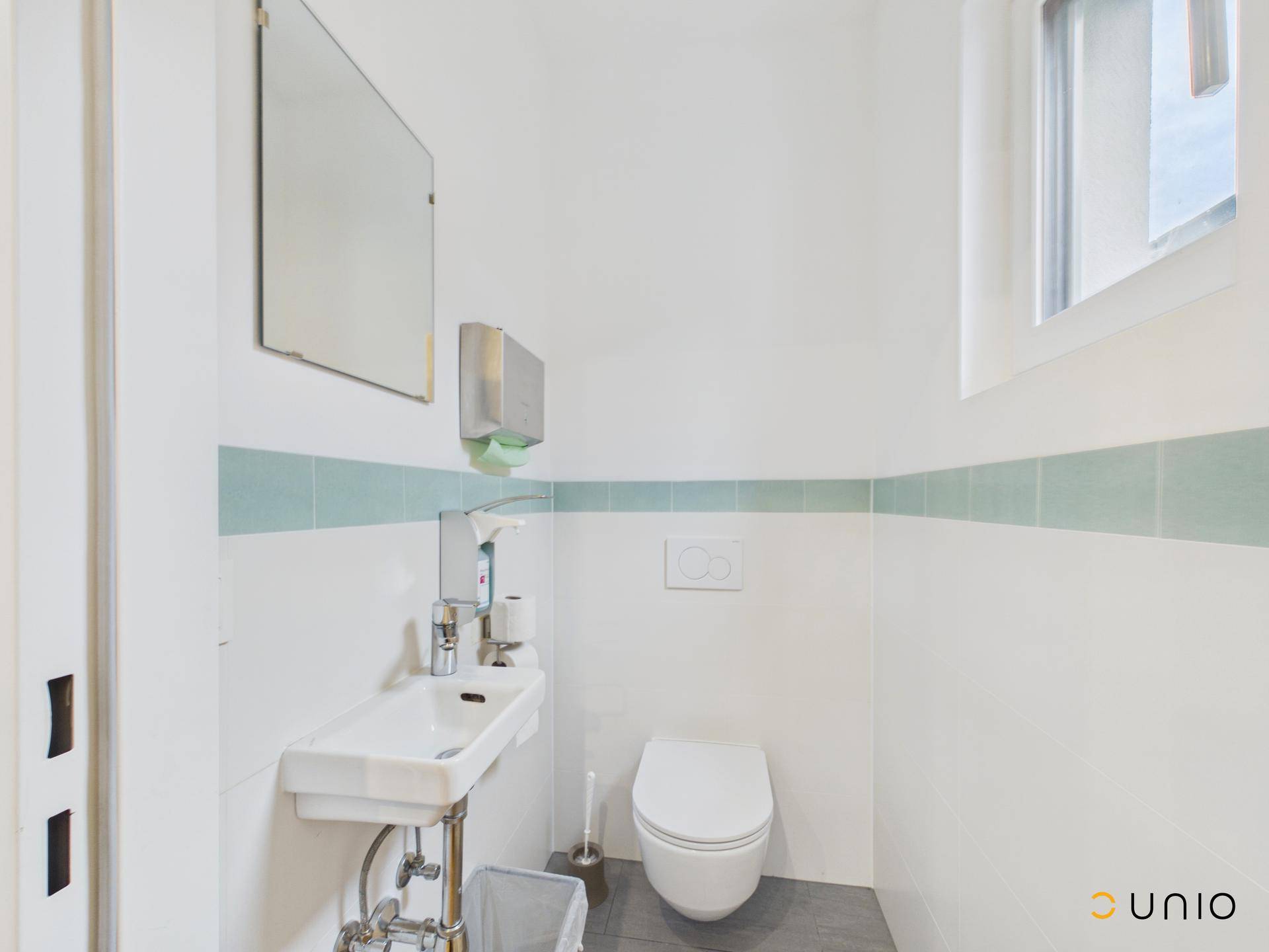 Helles WC mit wandhängender Toilette, kleinem Waschbecken und Fenster.