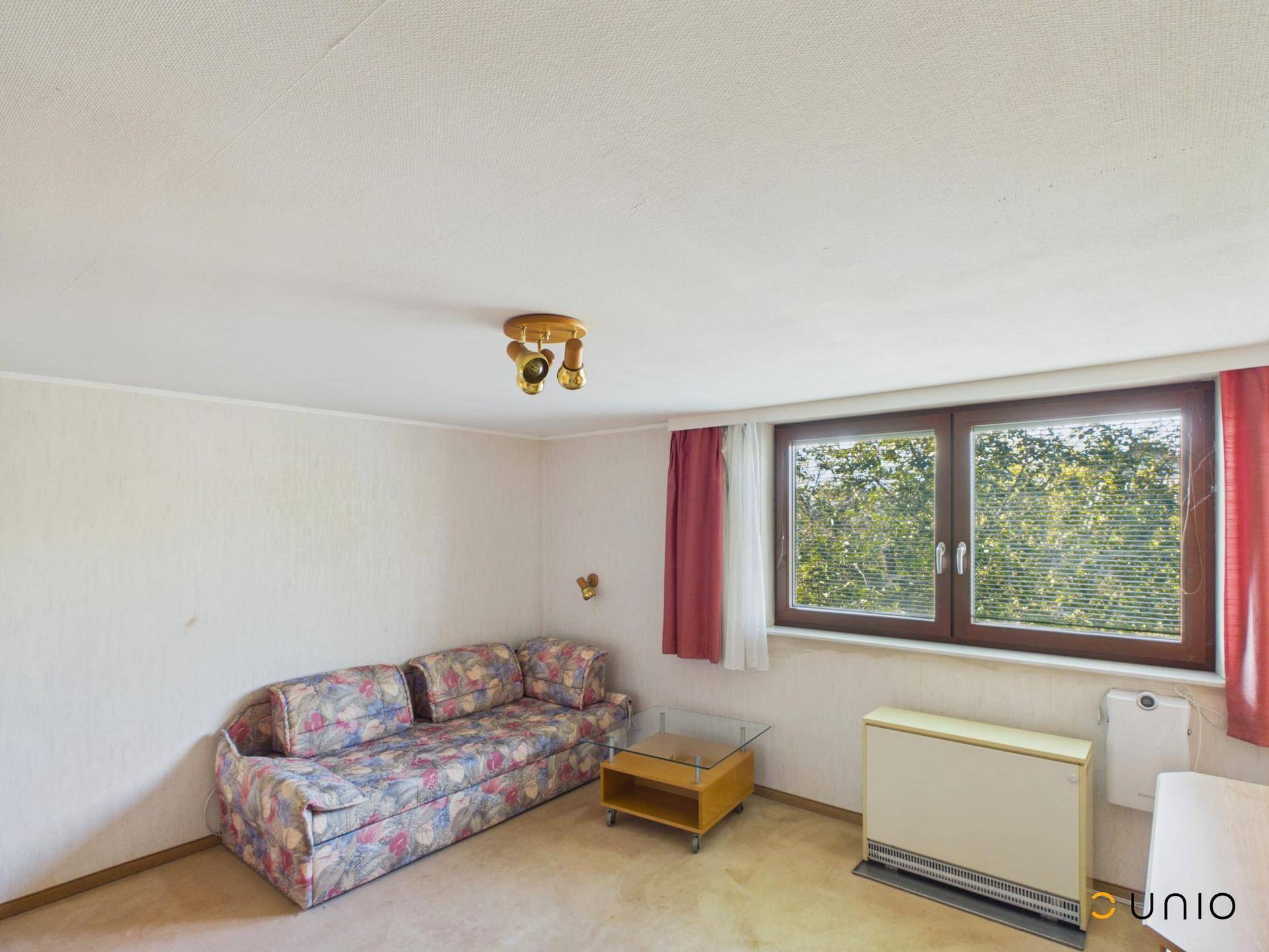 Wohnzimmer mit gemustertem Sofa, kleinem Tisch und Fenster mit Blick ins Grüne.