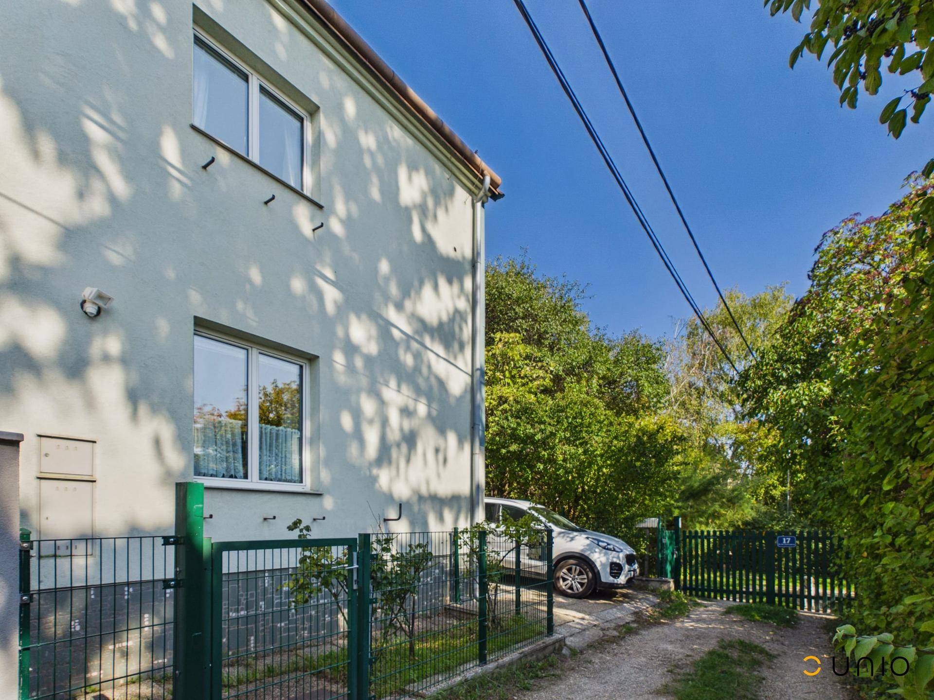 Nahaufnahme der Hausfassade mit Fenstern, grünem Zaun und üppiger Vegetation im Vorgarten.