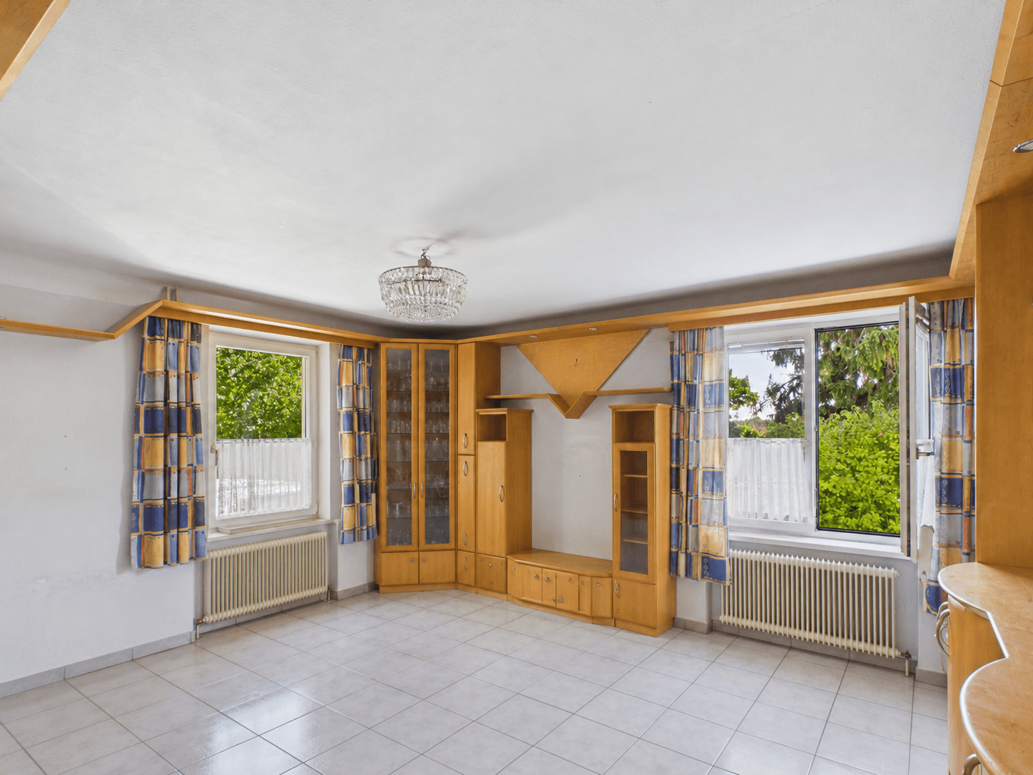 Geräumiges Wohnzimmer mit Fliesenboden, Einbauschränken und Blick ins Grüne durch die Fenster.