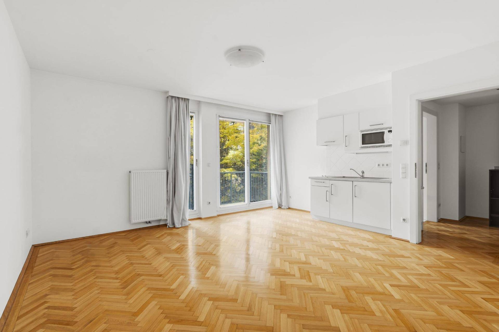 Leerer Wohnbereich mit Fischgrätparkett, Kochnische und großen Fenstern zum Balkon.