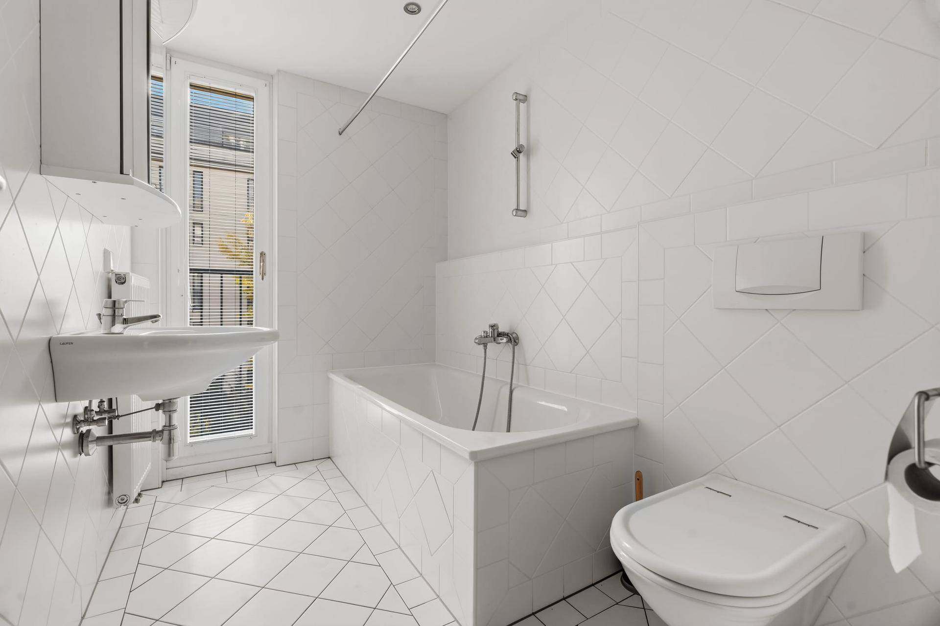 Helles Badezimmer mit weißen Fliesen, Badewanne, Waschbecken und Fenster.