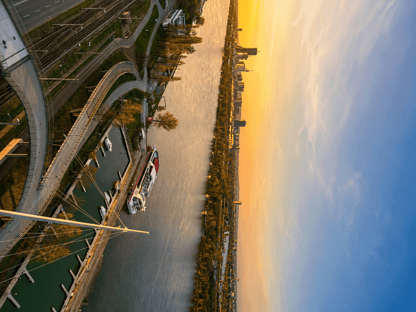 Luftaufnahme eines Flusses mit Yachthafen, Stadtpanorama und einer beeindruckenden Himmelsfärbung bei Sonnenuntergang.