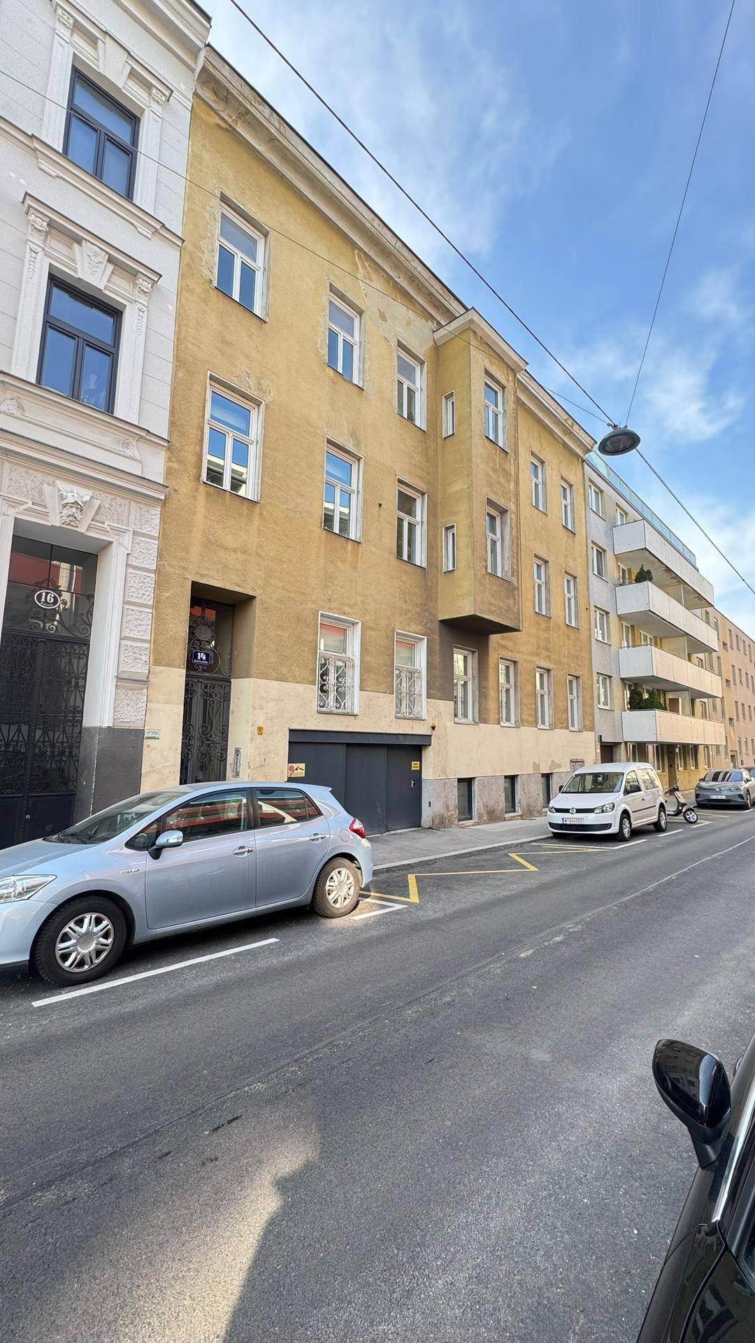 Stadtansicht einer Wohnstraße mit geparkten Autos und einem Mehrfamilienhaus mit Balkonen.