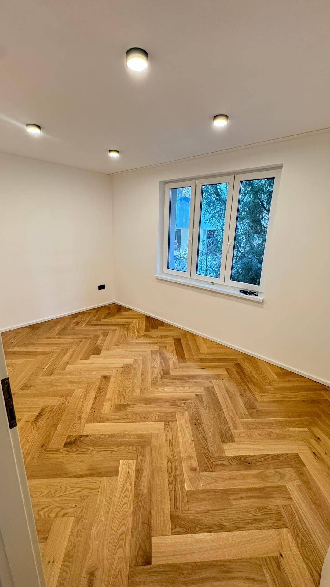Heller, leerer Raum mit Fischgrätparkettboden und großem Fenster, ideal für Schlaf- oder Arbeitszimmer.